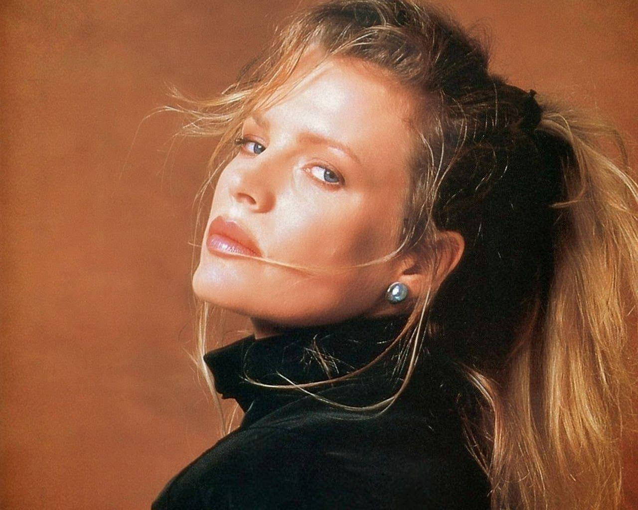 Kim Basinger Artistic Portrait 壁紙 — 一位金髮高馬尾的女性，在溫暖的棕色背景下，穿著黑色高領毛衣和珍珠耳環，若有所思地凝視著。