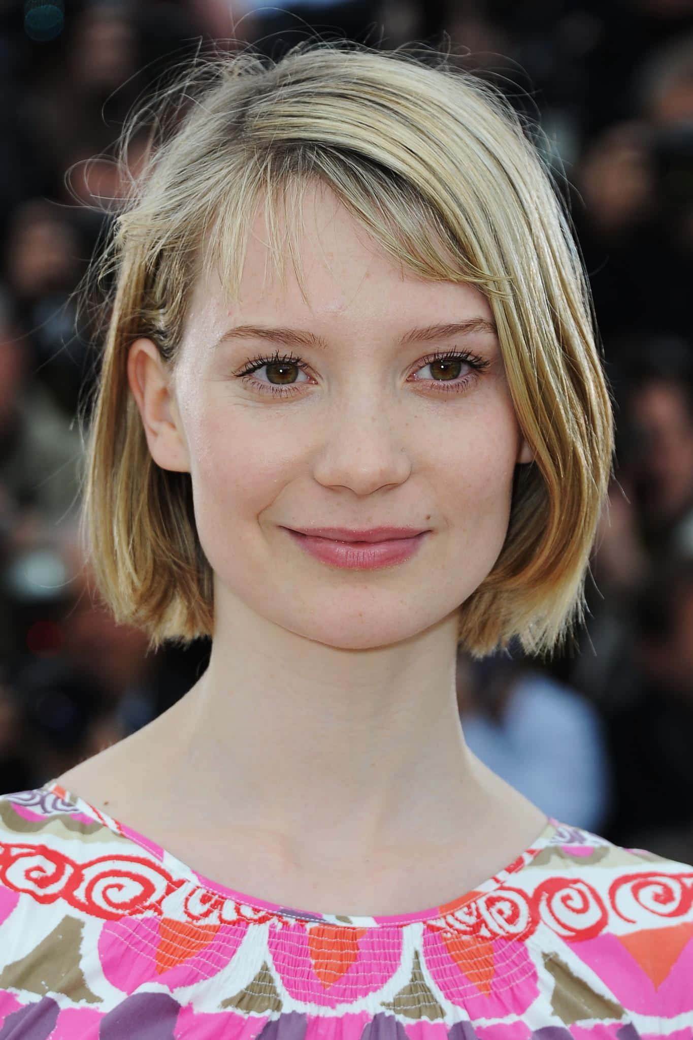 Mia Wasikowska 桌布 壁紙 — 一位金髮鮑伯頭女性嘅特寫肖像，帶住溫柔嘅笑容，身穿色彩繽紛嘅圖案上衣。
