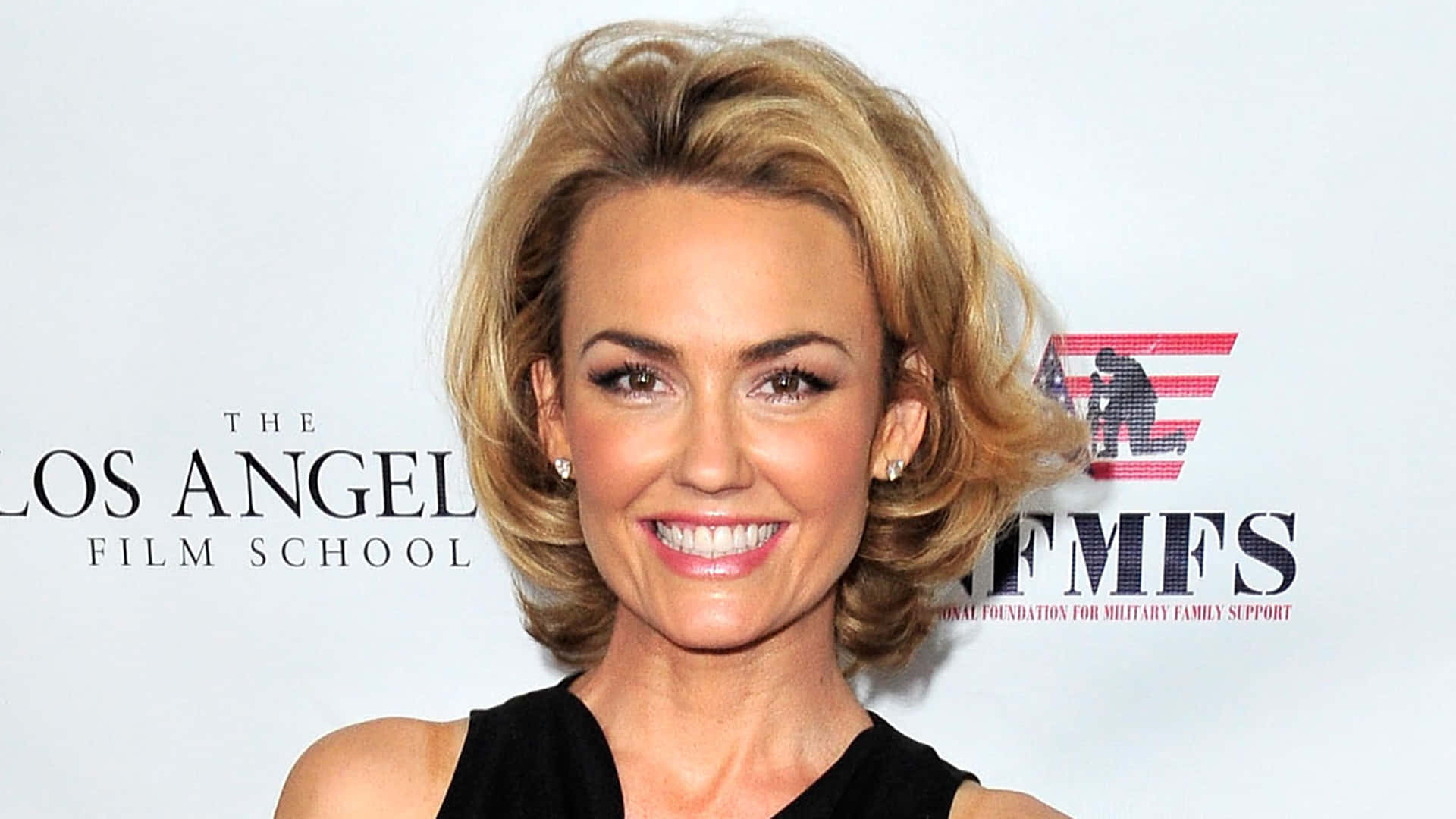 Kelly Carlson 桌布 壁紙 — 一位金髮黑裙的微笑女性在洛杉磯電影學院的活動中，於品牌背景前擺姿勢。