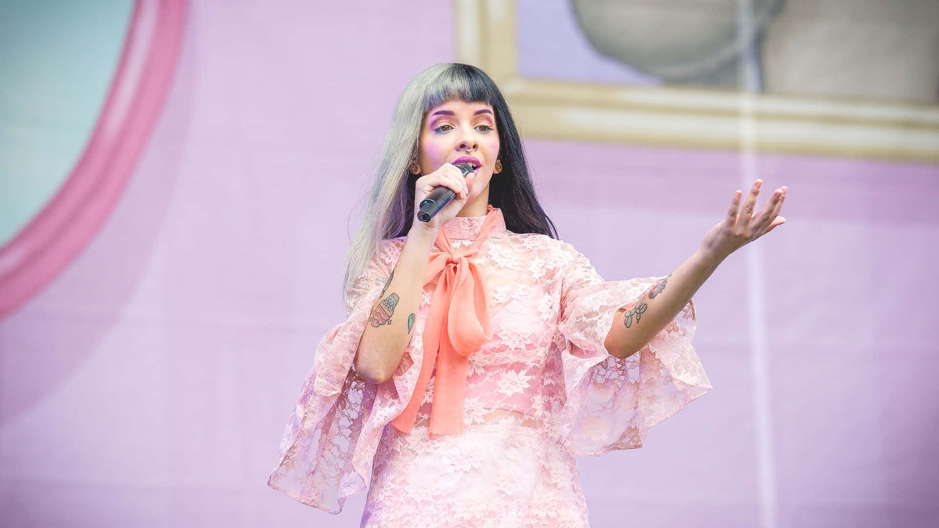 Melanie Martinez 桌布 壁紙 — 一位銀髮紋身女歌手身穿粉紅色蕾絲服裝在舞台上手持麥克風表演。
