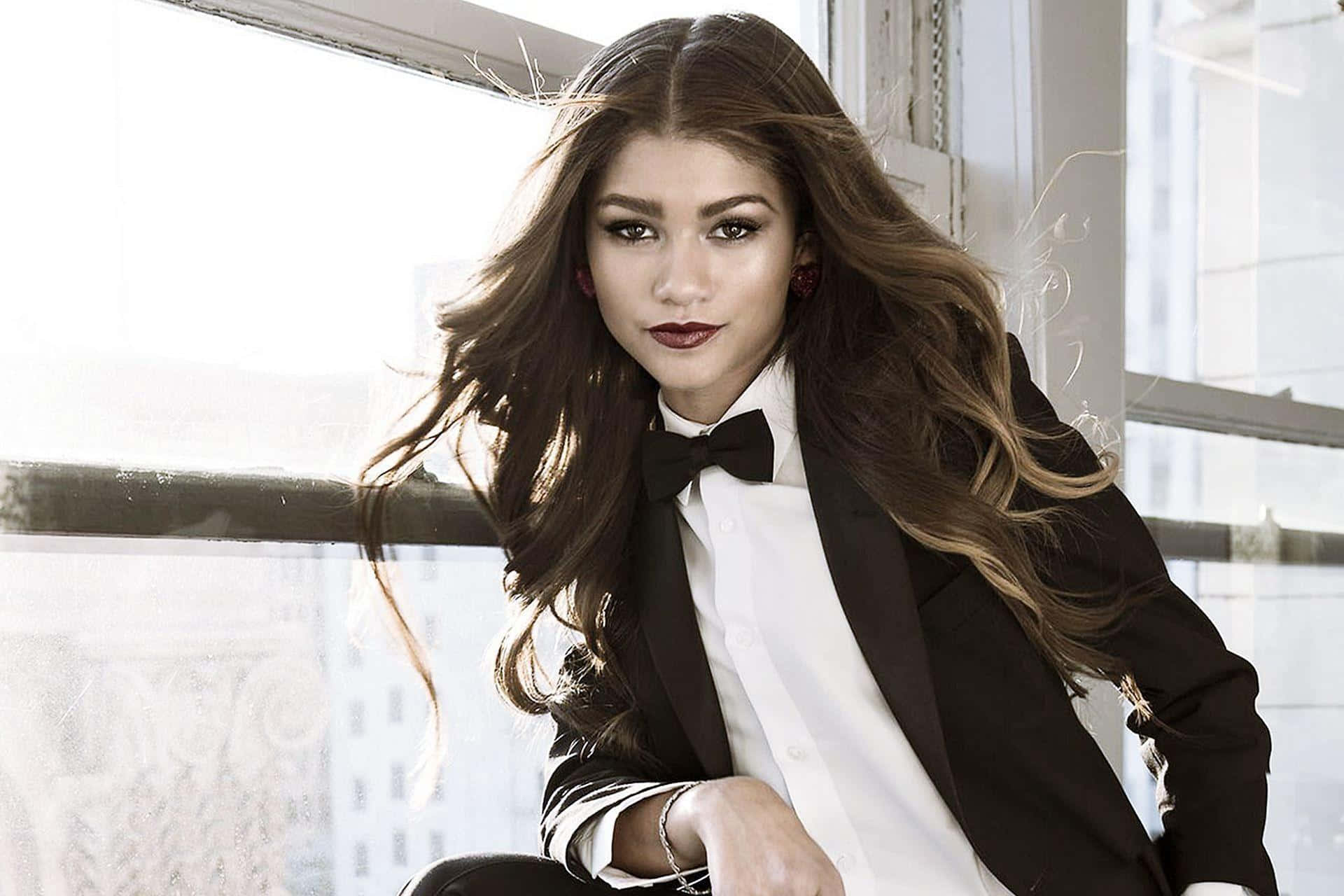 Zendaya 桌布 壁紙 — 一位長捲髮女性穿著黑色燕尾服和領結，在明亮的窗邊擺姿勢，身後可見城市建築。