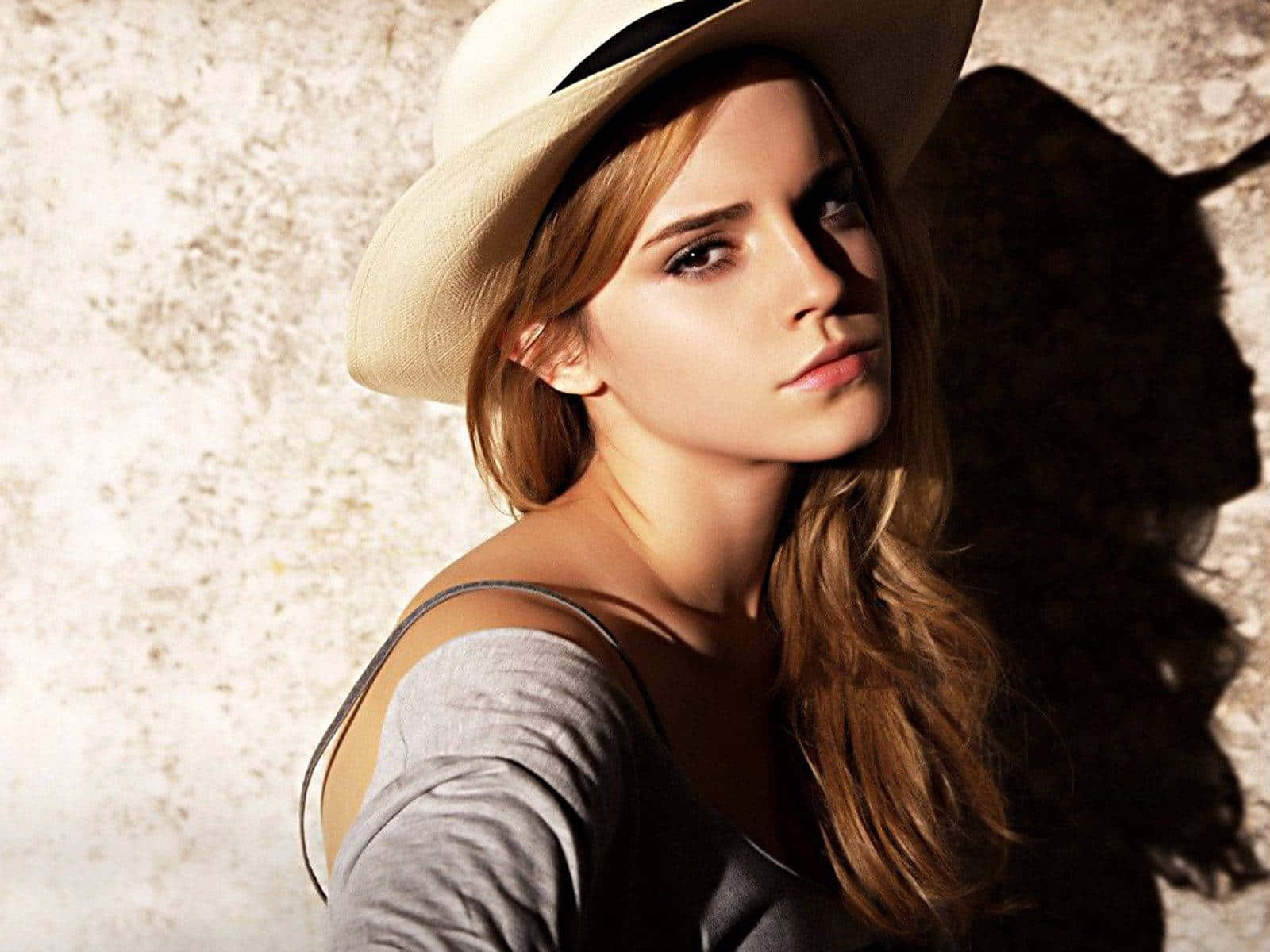 Emma Watson 桌布 壁紙 — 一位長棕色頭髮的女性戴著米色帽子和灰色上衣，靠著紋理牆壁，戲劇性的燈光投射出強烈的陰影。