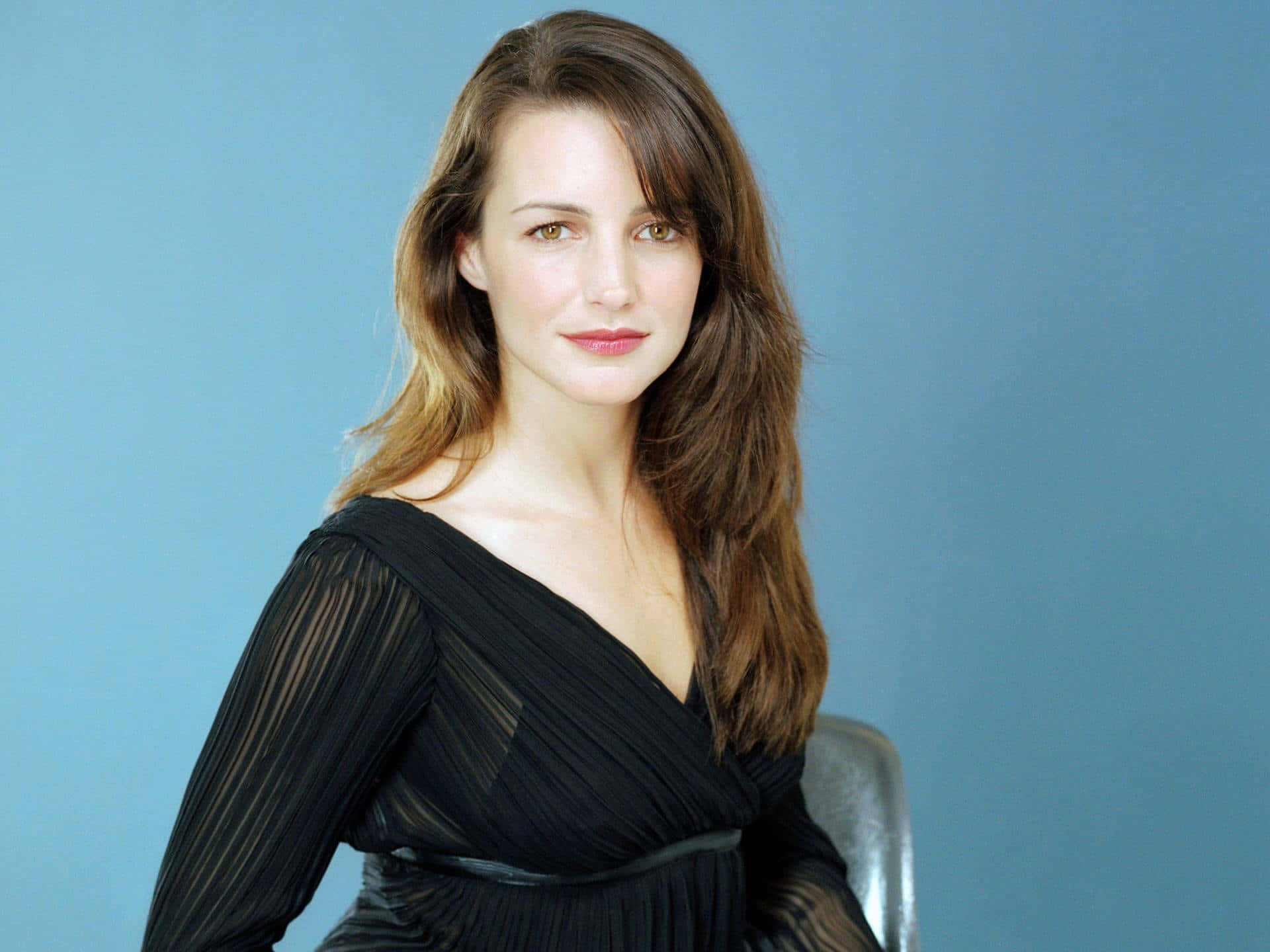 kristin davis 桌布 壁紙 — 一位長棕色頭髮的女性穿著黑色百褶連衣裙，背景為純藍色攝影棚背景。
