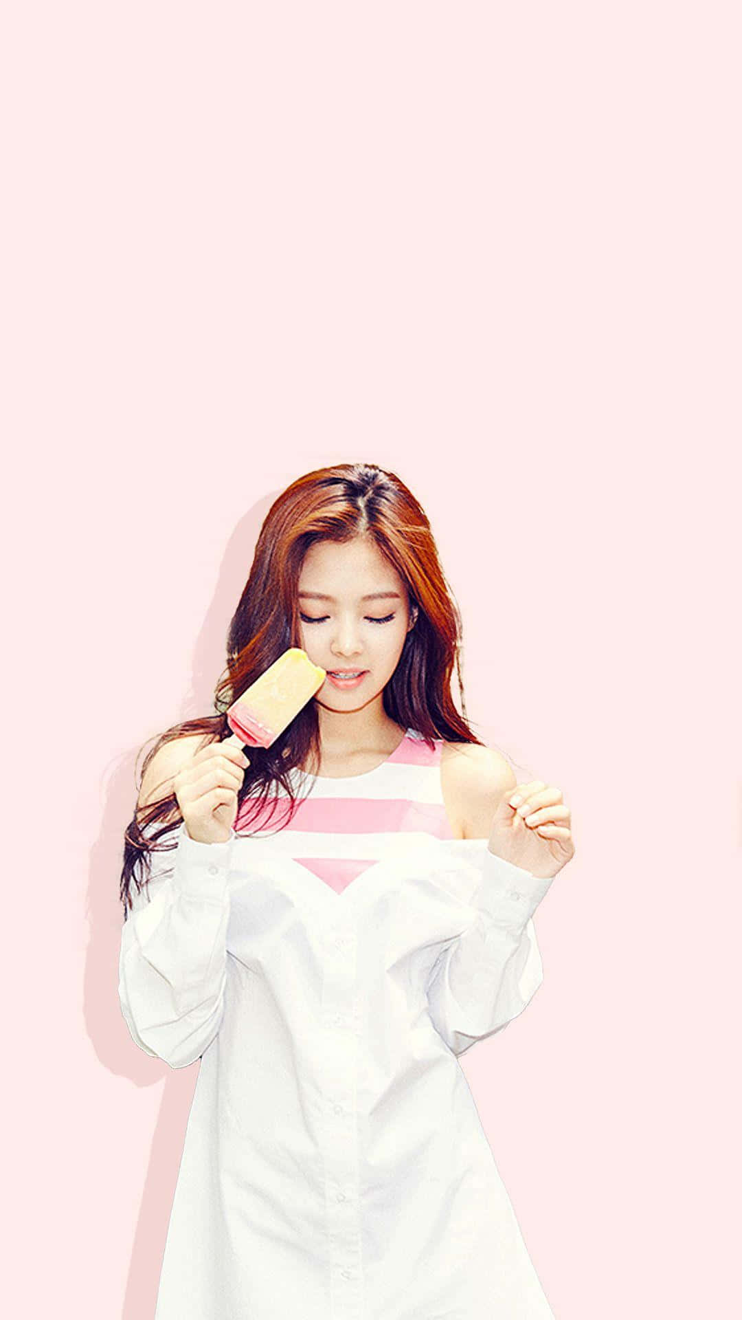 Jennie Kim 桌布 壁紙 — 一位長棕色頭髮的年輕女性在柔和的粉紅色背景前享用冰棒。