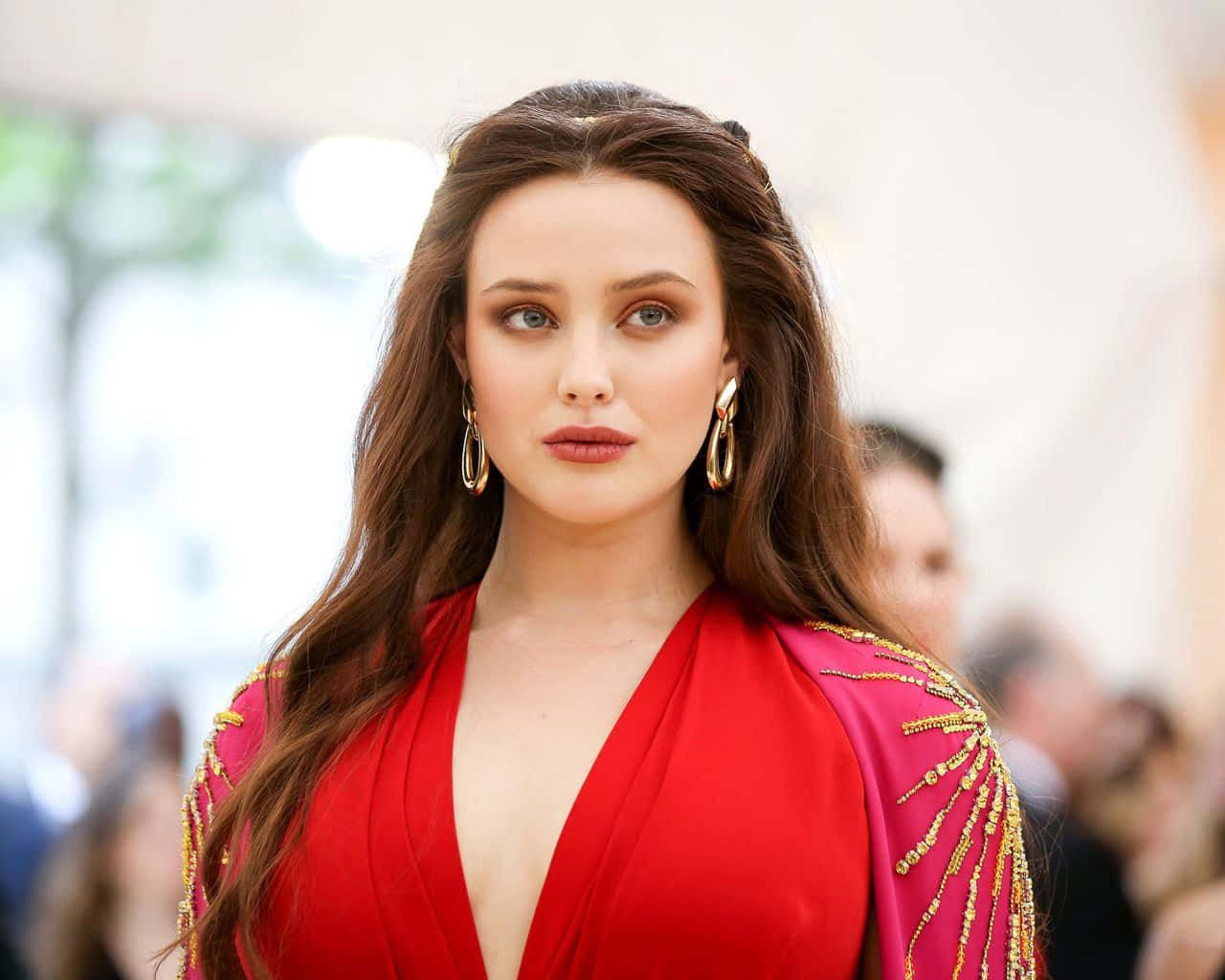 Met Gala 桌布 壁紙 — 一位長棕髮女性穿著鮮紅色連衣裙，肩部飾有金色珠飾，戴著大圈耳環。