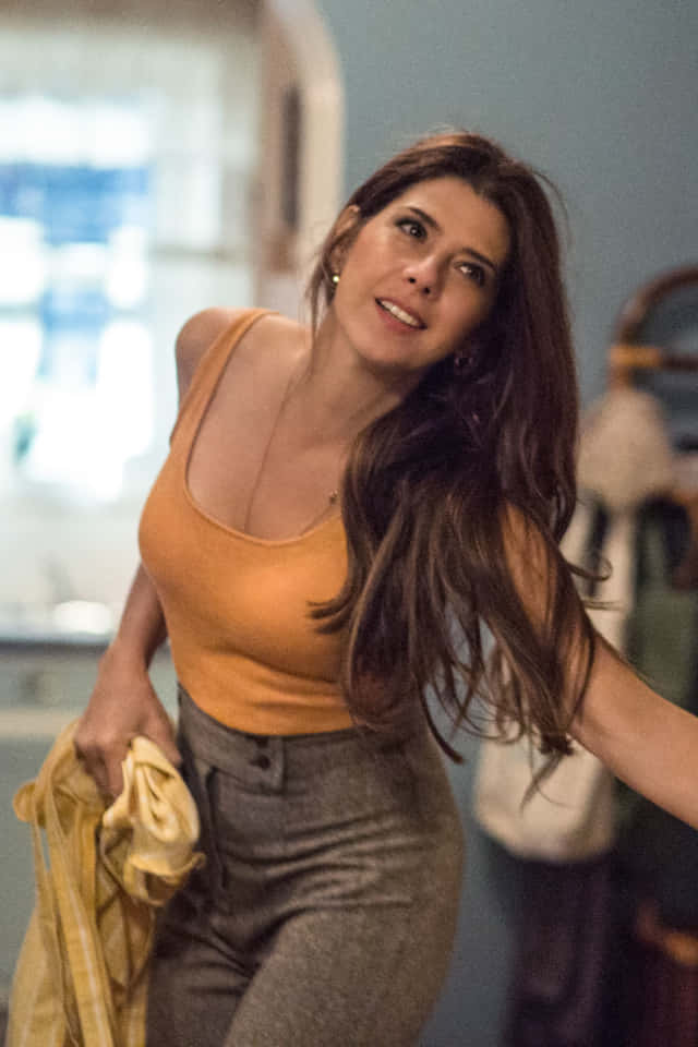 Aunt May 桌布 壁紙 — 一位長棕髮的女性在柔和光線的室內空間微笑著，手持一件黃色衣物。