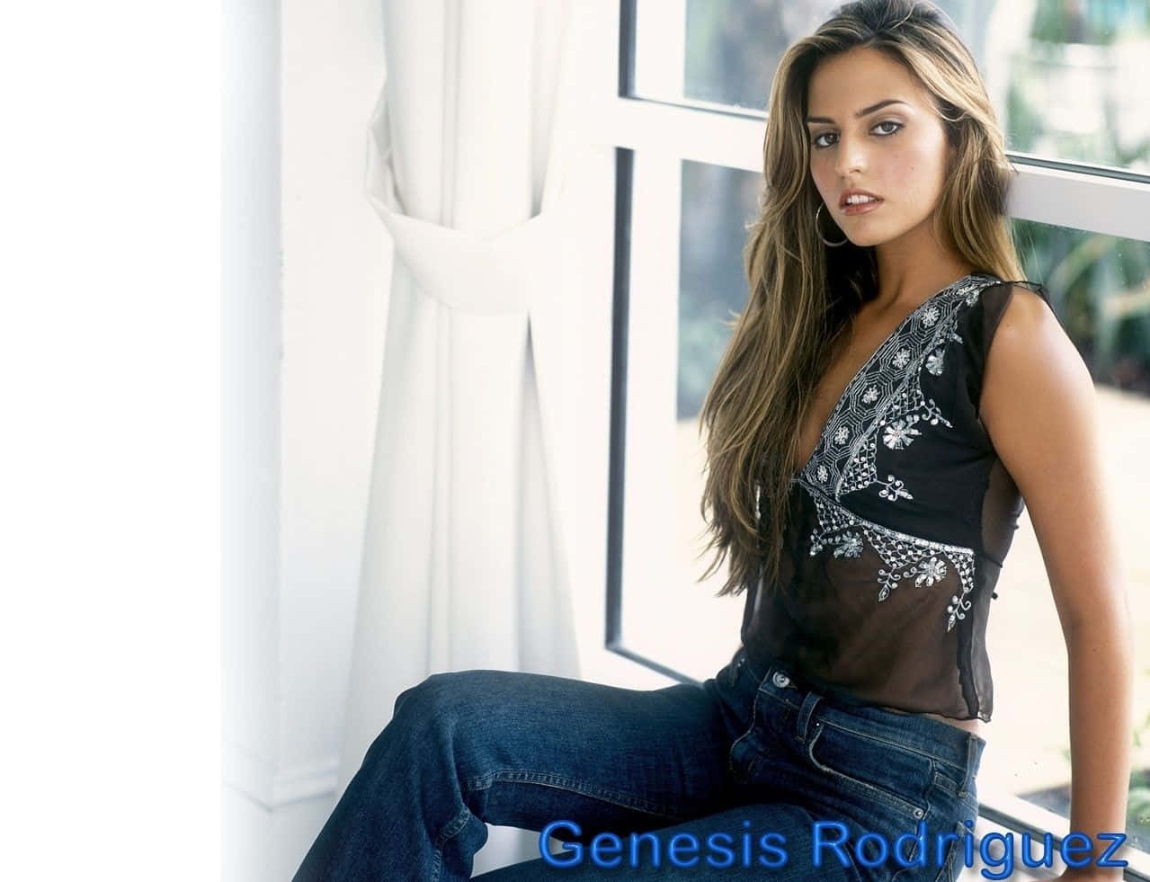 Genesis Rodriguez 桌布 壁紙 — 一位長棕髮的女性坐在明亮的窗邊，身穿黑色刺繡上衣和牛仔褲。