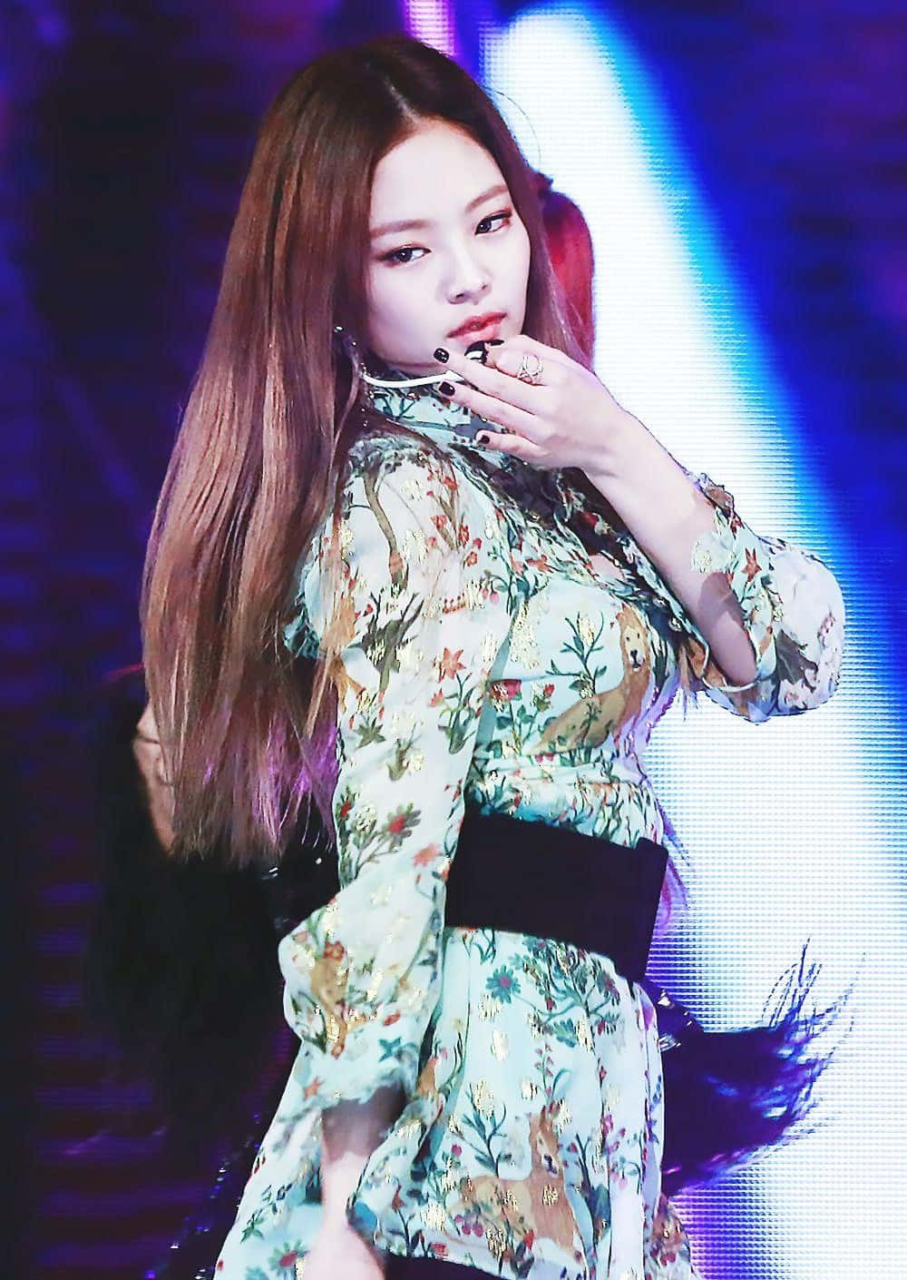 Jennie Kim 桌布 壁紙 — 一位長棕髮的女表演者穿著花卉圖案連衣裙，在充滿活力的藍色舞台燈光下自信地擺姿勢。