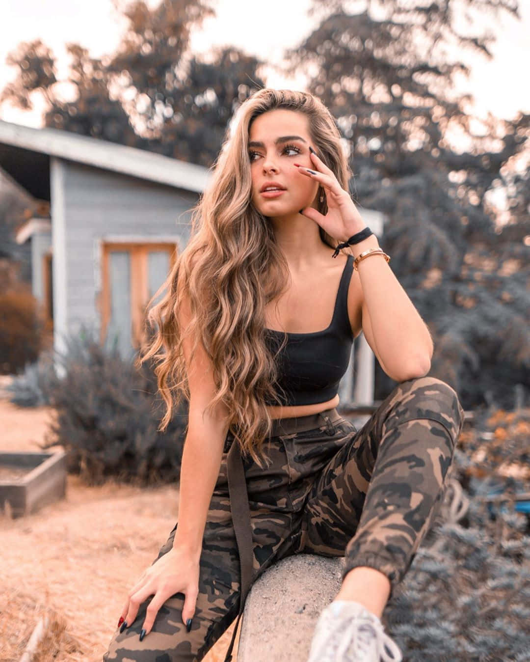 Addison Rae 桌布 壁紙 — 一位長波浪髮的女性，穿著迷彩褲和黑色短上衣，隨意地坐在岩石上，背景是質樸的房子。
