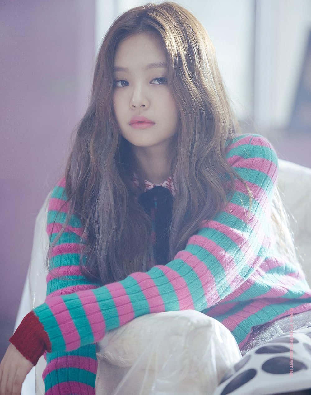 Jennie Kim 桌布 壁紙 — 一位長波浪髮的年輕女性穿著充滿活力的粉紅和藍綠色條紋毛衣，坐在室內，周圍是柔和的紫色燈光。