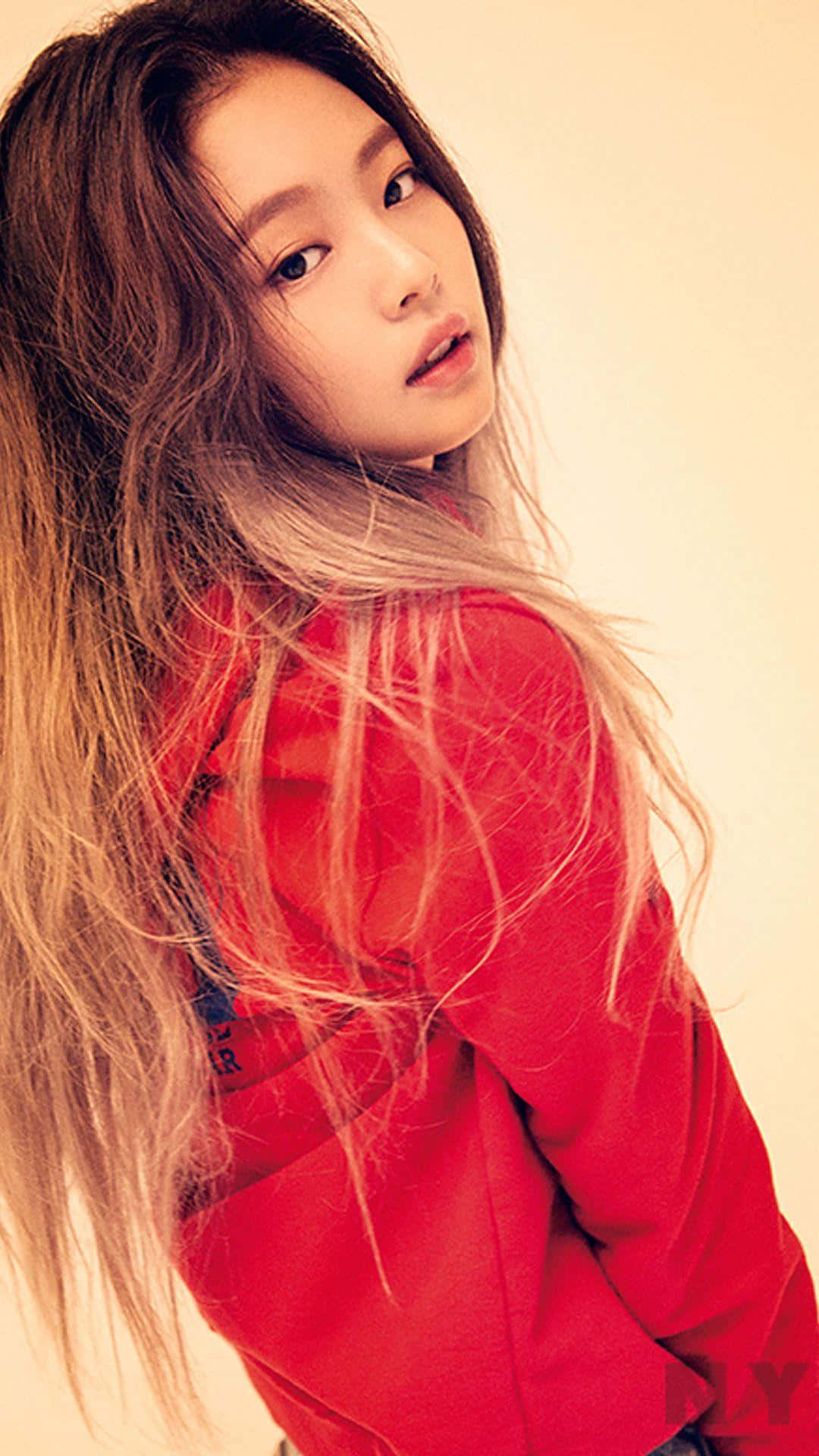 Jennie Kim 桌布 壁紙 — 一位長波浪髮的年輕女性穿著紅色連帽衫，在溫暖的米色背景上轉頭越過肩膀。