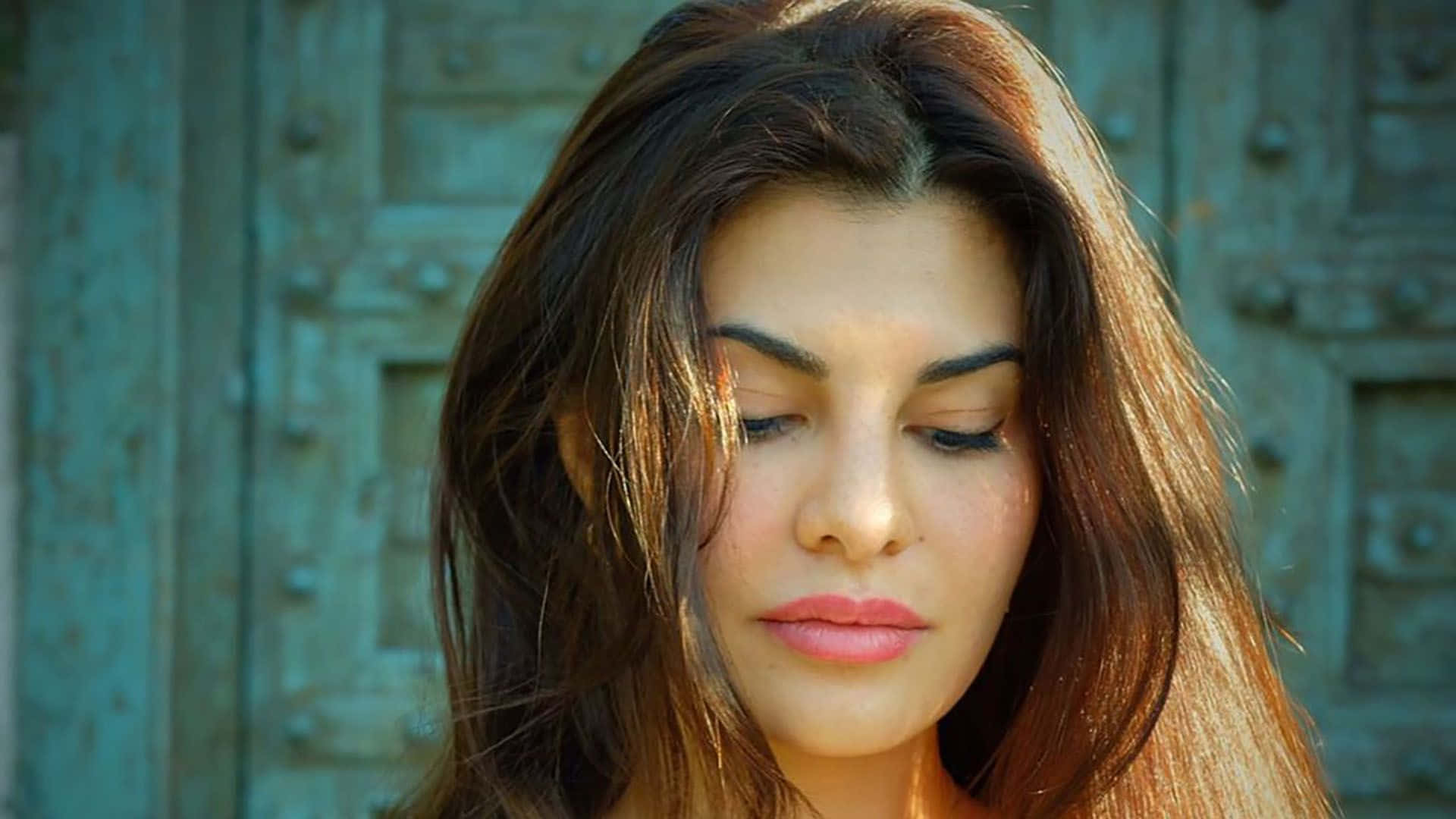 Jacqueline Fernandez 桌布 壁紙 — 一位長著棕色長髮、閉著眼睛的女性站在一扇飽經風霜的藍綠色門前，沐浴在溫暖的陽光下。