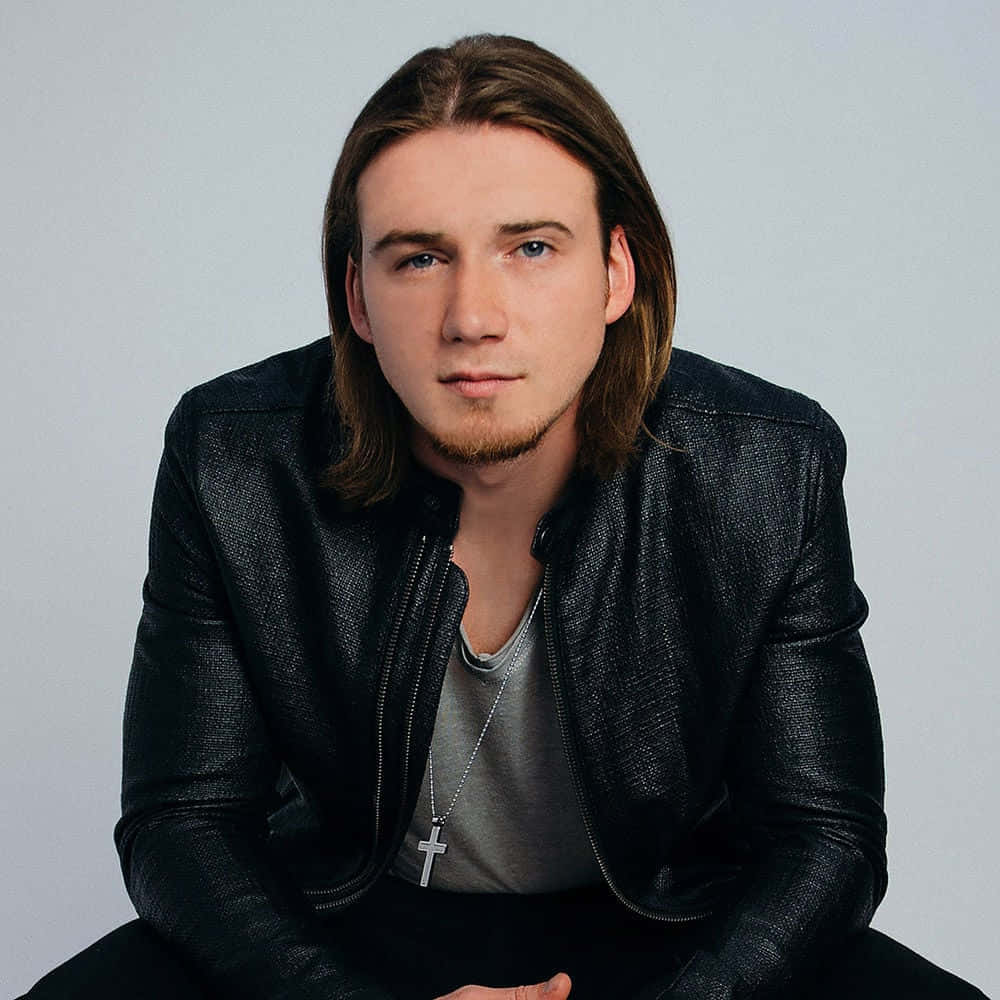 Morgan Wallen 桌布 壁紙 — 一位長著棕色長髮和藍色眼睛的年輕男子，穿著黑色皮夾克，內搭灰色襯衫，戴著十字架項鍊，背景是純淺灰色。