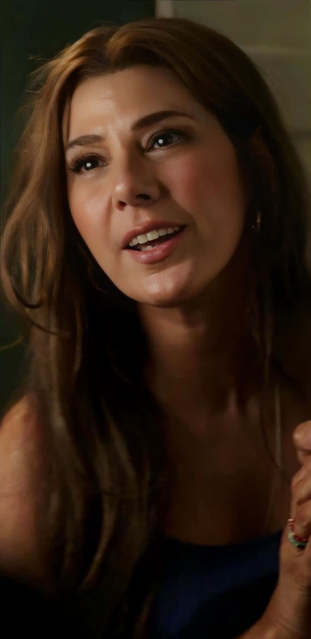 Aunt May 桌布 壁紙 — 一位長著棕色長髮的女性在柔和昏暗的燈光下穿著藍色上衣溫暖地微笑。