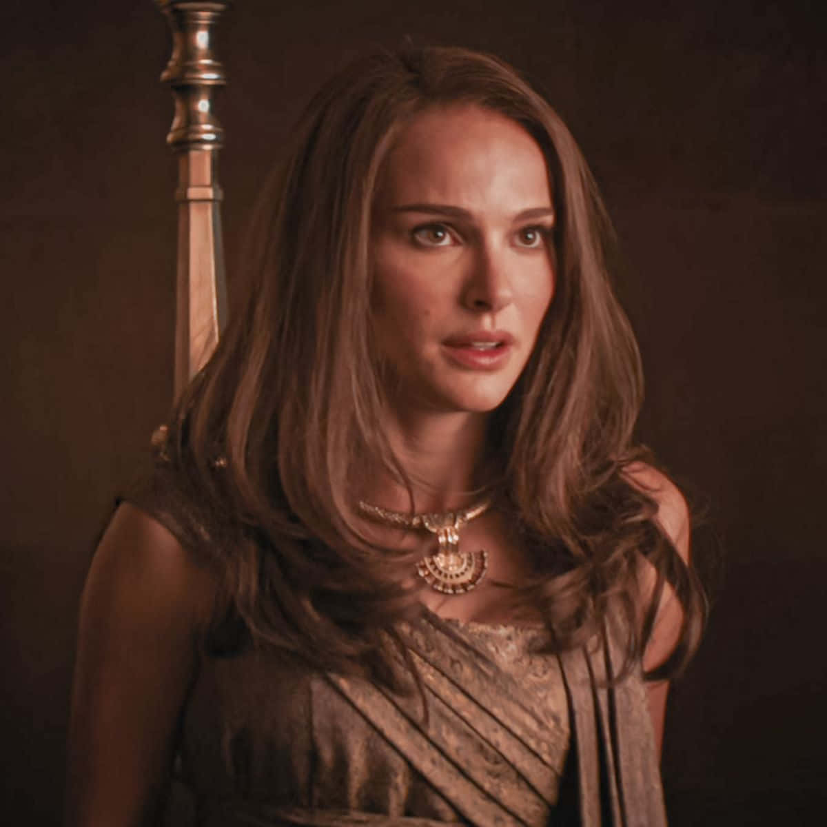 Jane Foster 桌布 壁紙 — 一位長著棕色長髮的女性戴著華麗的金項鍊和垂墜的服裝，站在一個高大的金屬桿旁，光線昏暗。