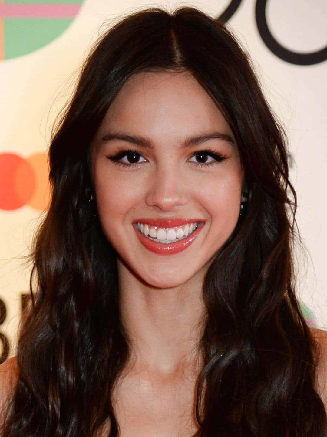 Olivia Rodrigo 桌布 壁紙 — 一位長著深色波浪長髮的耀眼女性，在色彩繽紛的抽象背景前燦爛微笑。