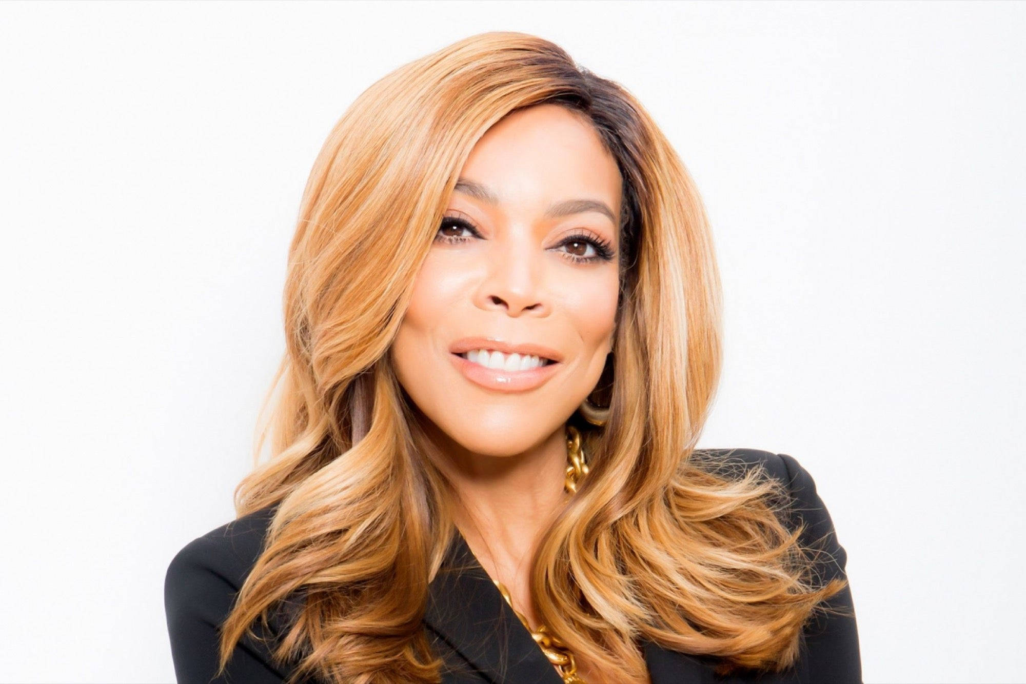 Wendy Williams Simple Hairstyle 壁紙 — 一位長著長波浪金髮的容光煥發的女性，在純白色背景前微笑，身穿黑色西裝外套和金色珠寶。