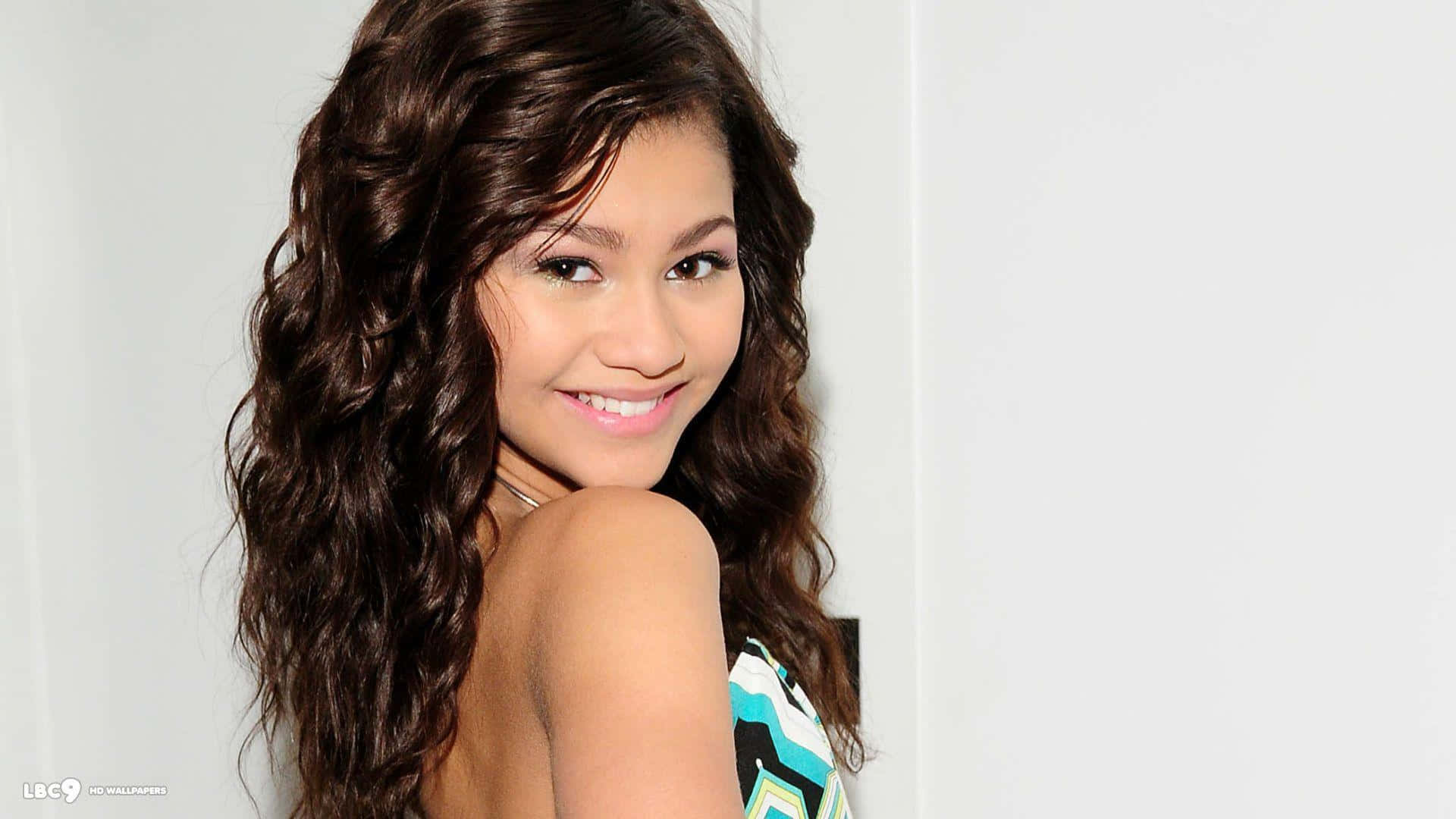 Zendaya 桌布 壁紙 — 一位長著長長波浪棕色頭髮的年輕女性，在純白牆壁前回頭微笑。