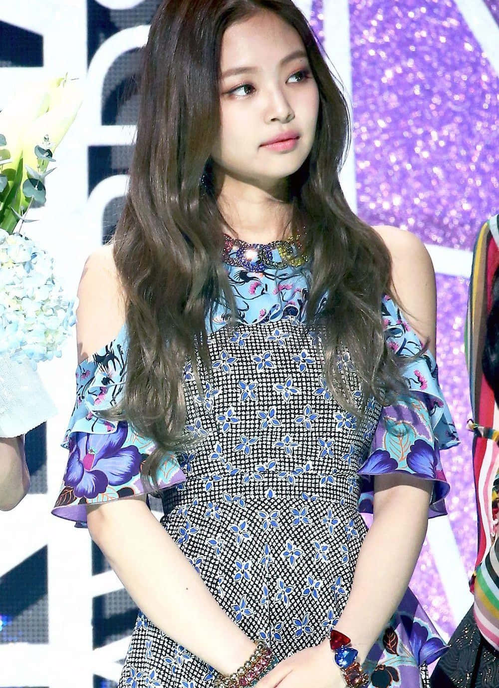 Jennie Kim 桌布 壁紙 — 一位長著長長波浪棕色頭髮的年輕女性，穿著荷葉邊袖子的花卉圖案連衣裙，背景是閃閃發光的紫色和白色。