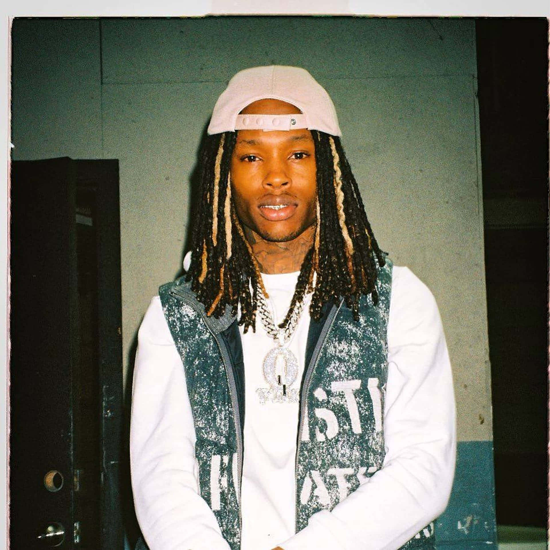 King Von 桌布 壁紙 — 一位長辮子、戴粉紅色帽子的年輕男子自信地站在燈光昏暗的室內空間，身穿閃亮背心和多層鏈條。