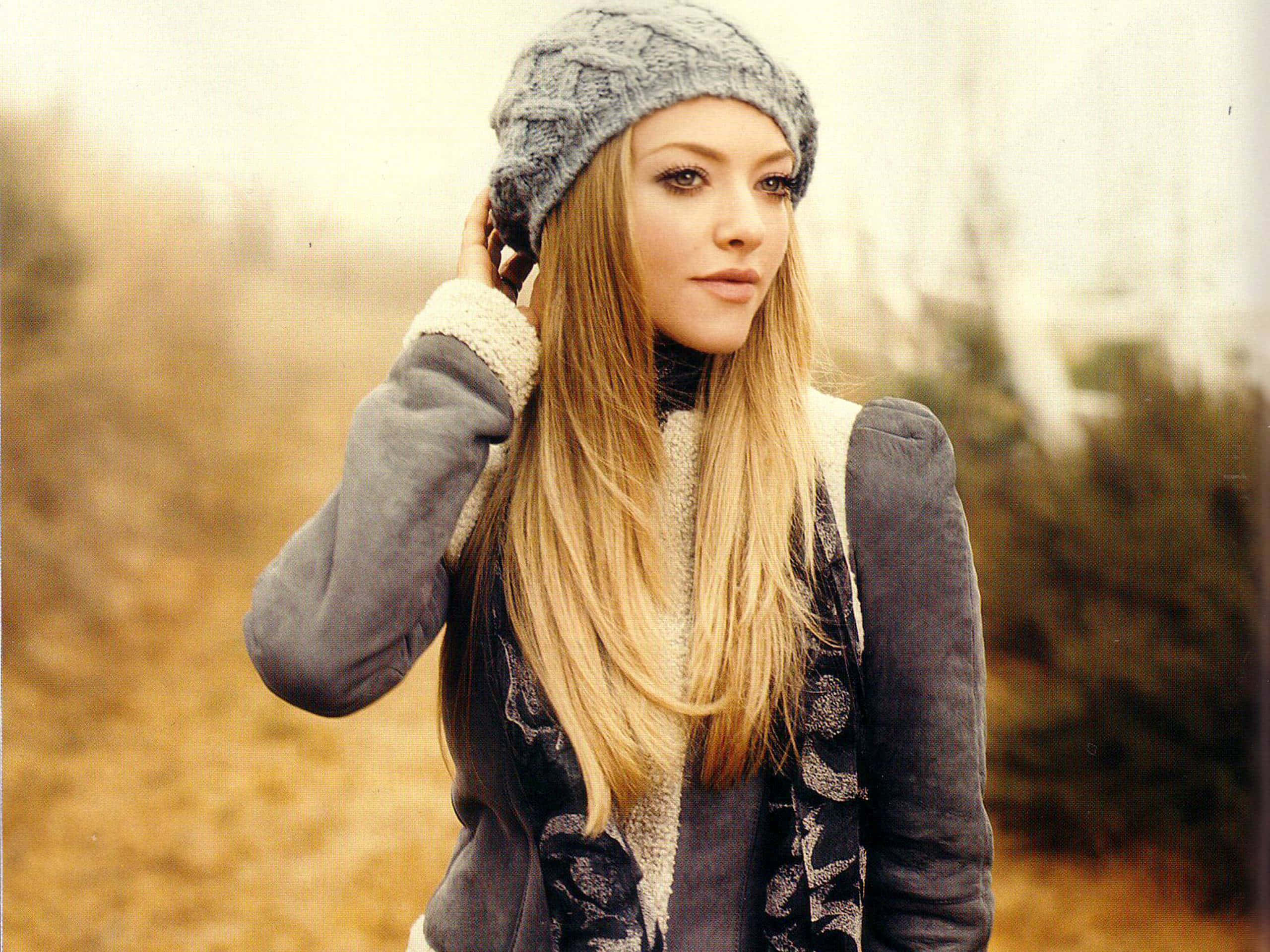 Amanda Seyfried 桌布 壁紙 — 一位長金髮的女性戴著灰色針織無邊帽和分層外套，站在戶外柔和的秋日環境中。