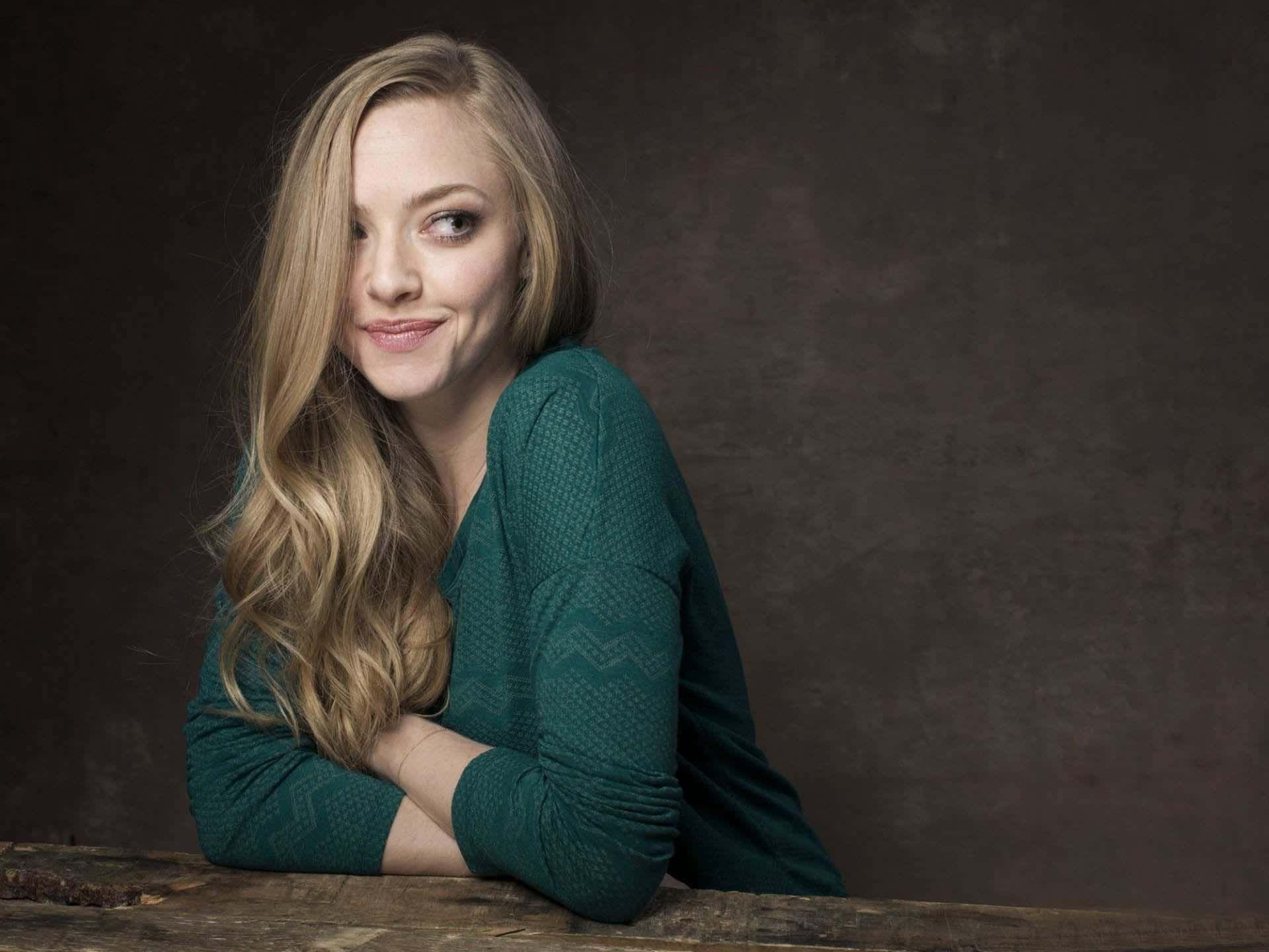 Amanda Seyfried 桌布 壁紙 — 一位長金髮的女性穿著青綠色質感毛衣，靠著木質表面，背景為深棕色。
