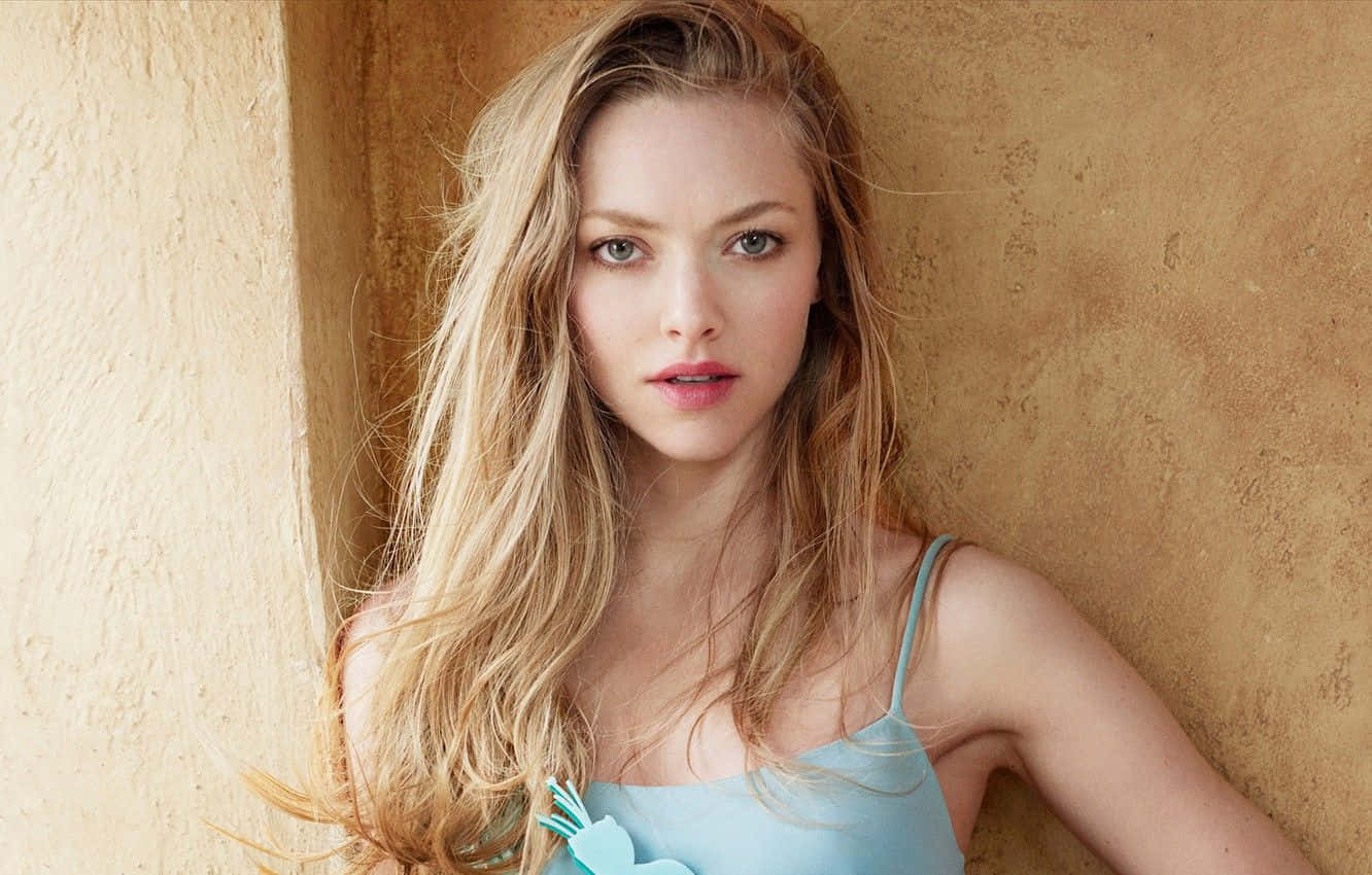 Amanda Seyfried 桌布 壁紙 — 一位長金髮的年輕女性靠著帶有紋理的米色牆壁，身穿淺藍色上衣。