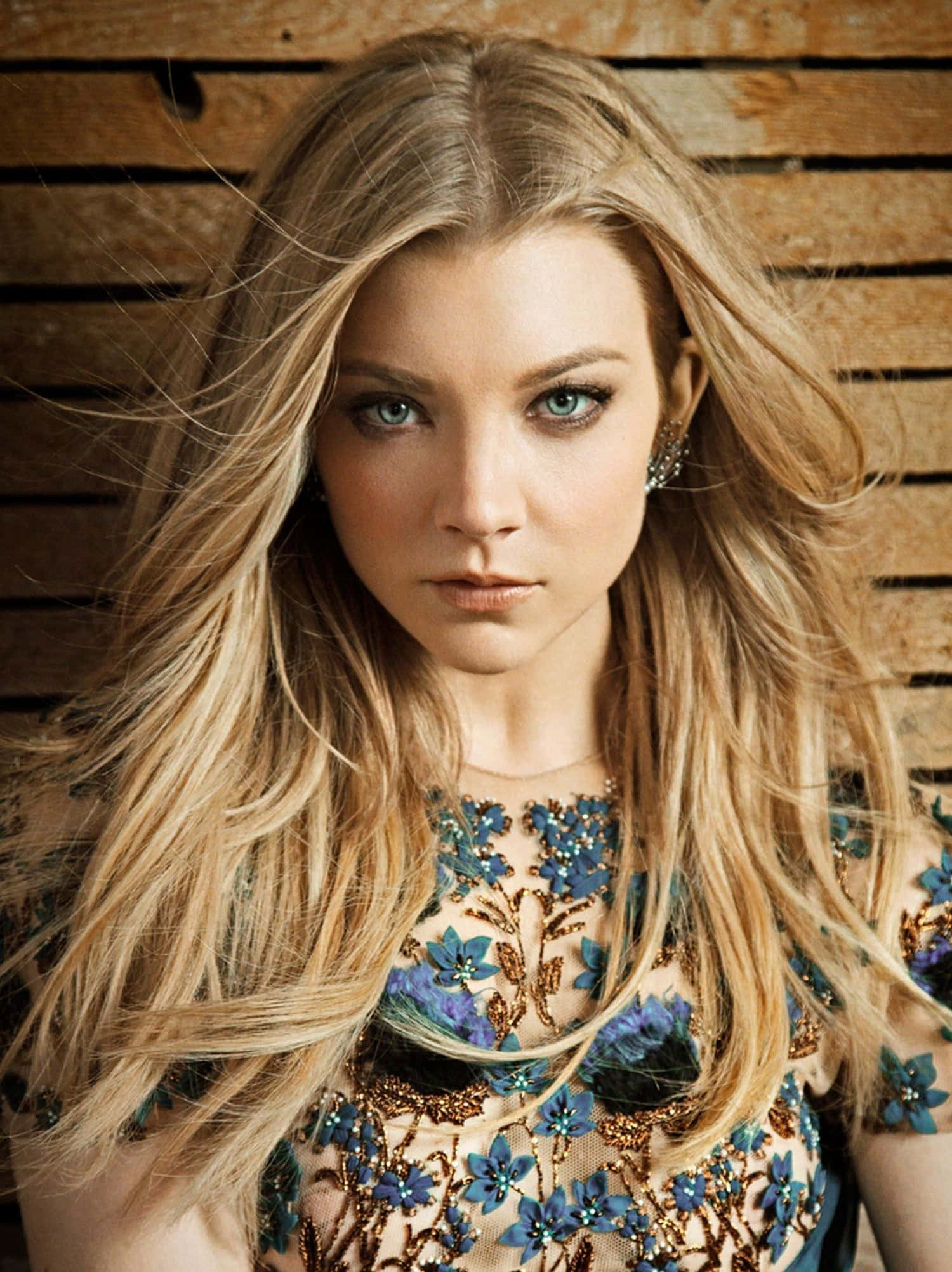 natalie dormer 桌布 壁紙 — 一位長金髮藍眼睛的女性，穿著花卉刺繡上衣，在木條背景前直視鏡頭。