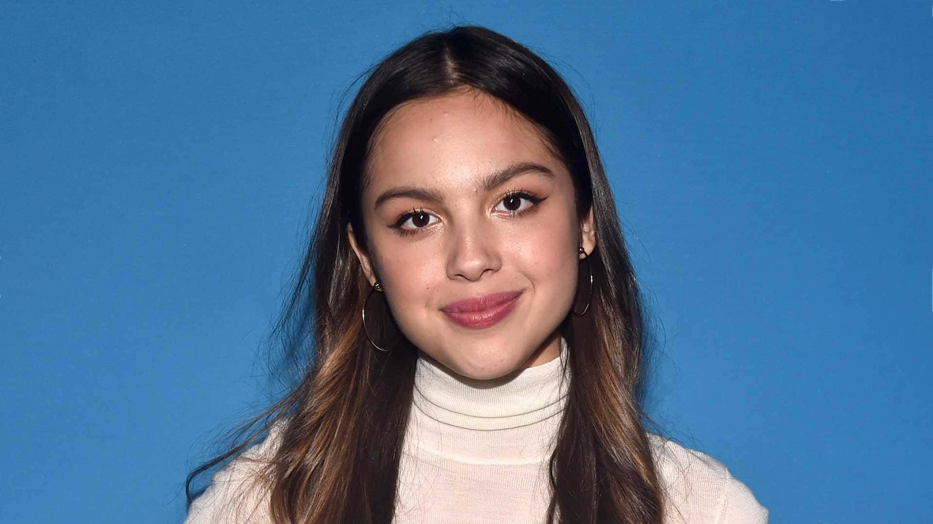 Olivia Rodrigo 桌布 壁紙 — 一位長長棕色頭髮的年輕女性，在純藍色背景前穿著奶油色高領毛衣，溫柔地微笑。