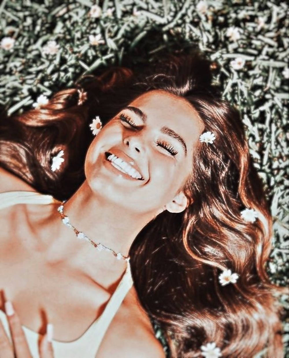 Smile 桌布 壁紙 — 一位長長棕色頭髮的快樂女性，躺在綠色草地和白色雛菊中微笑，穿著白色上衣和花卉項鍊。