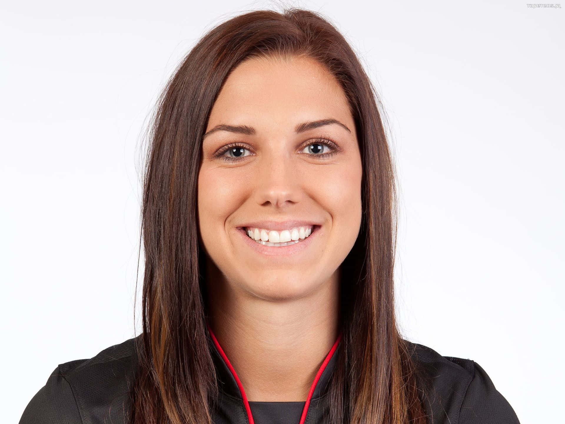 Alex Morgan 桌布 壁紙 — 一位長長棕色頭髮的開朗女性，穿著帶有紅色飾邊的黑色外套，在純白色背景前燦爛微笑。