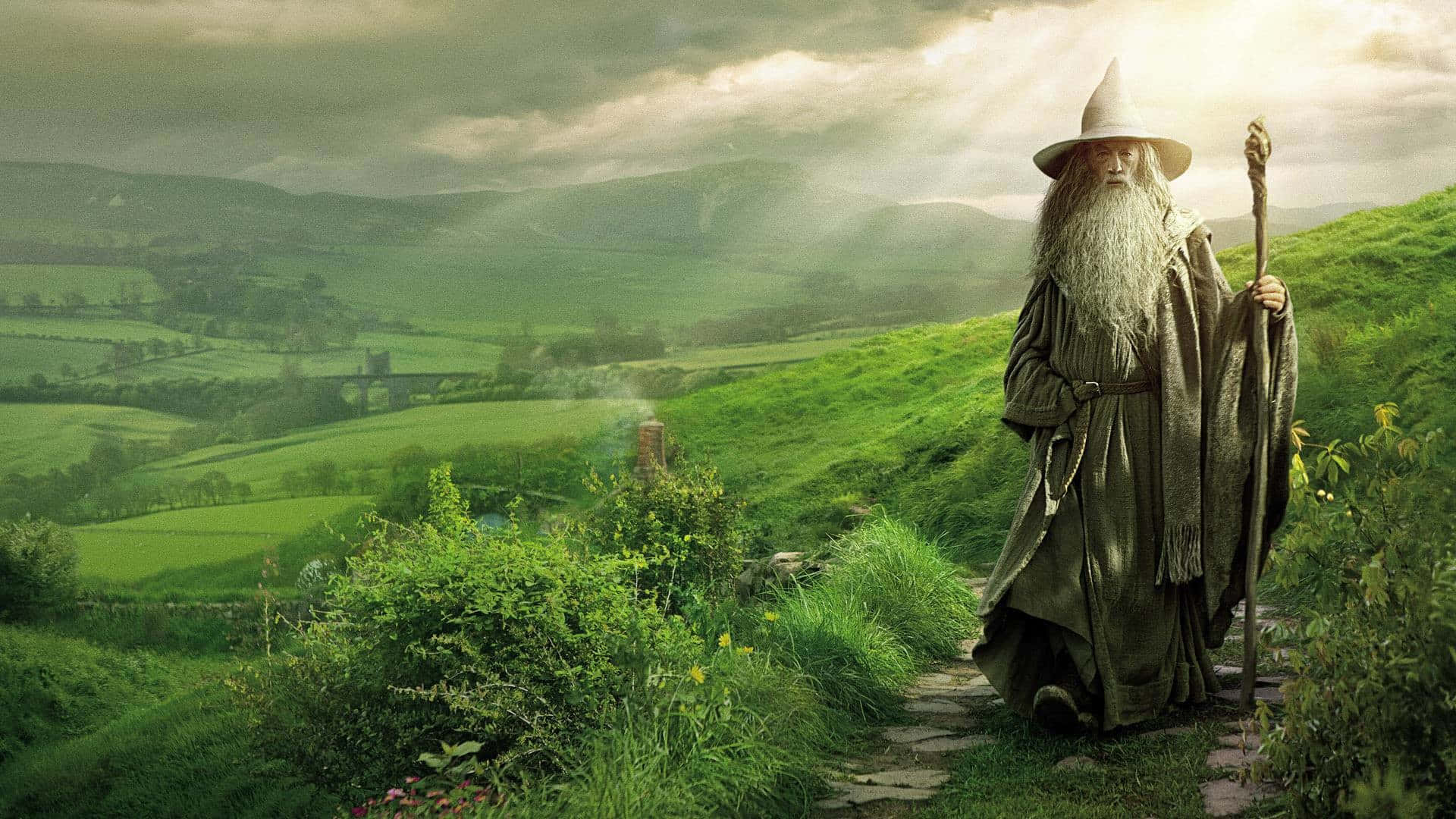 the hobbit 桌布 壁紙 — 一位長長白鬍鬚、戴尖帽的老巫師站在石徑上，俯瞰雲層間陽光穿透的陰天下一片翠綠的山谷。