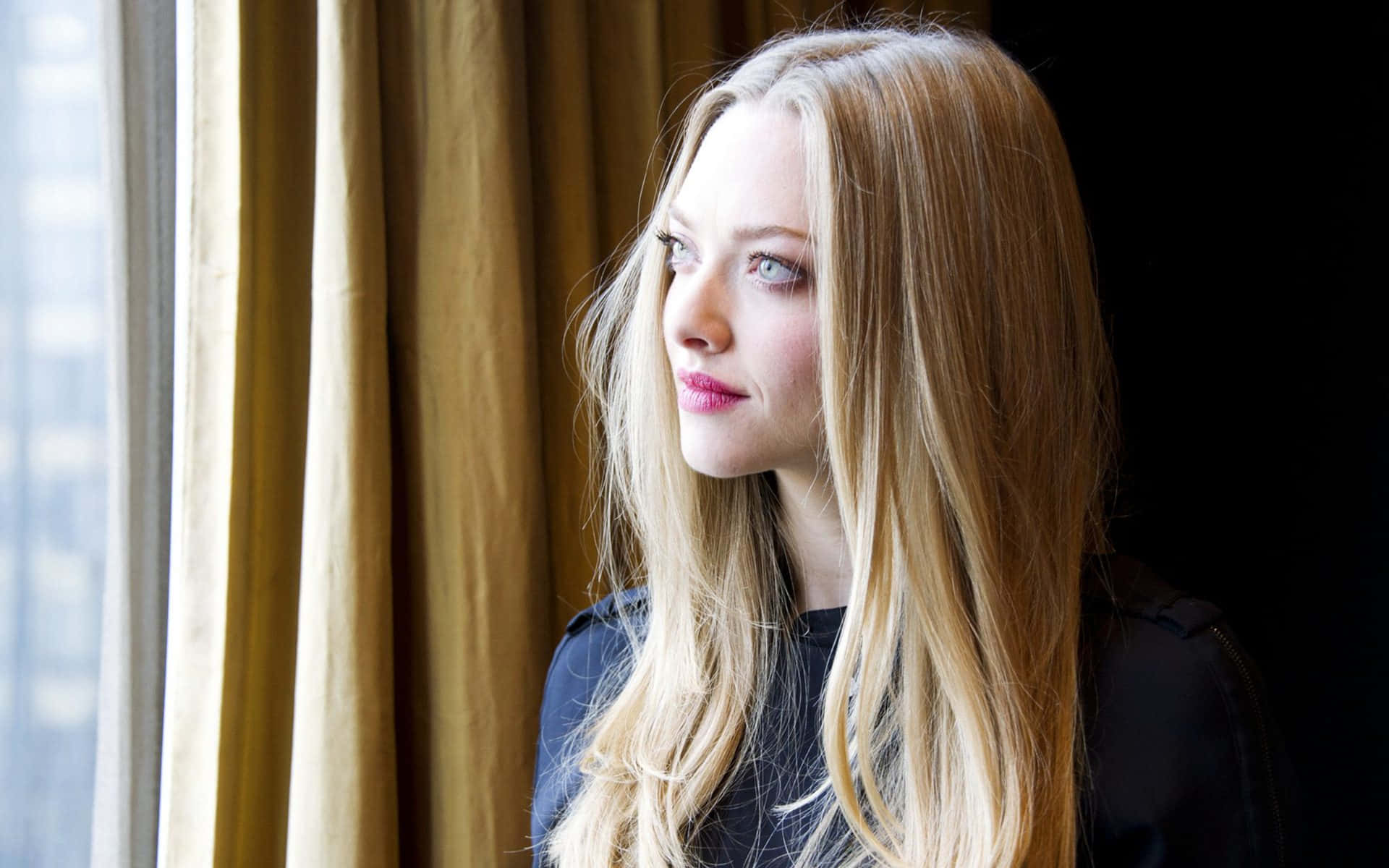 Amanda Seyfried 桌布 壁紙 — 一位長長金髮的女性，若有所思地望著被金色窗簾框住的窗外。