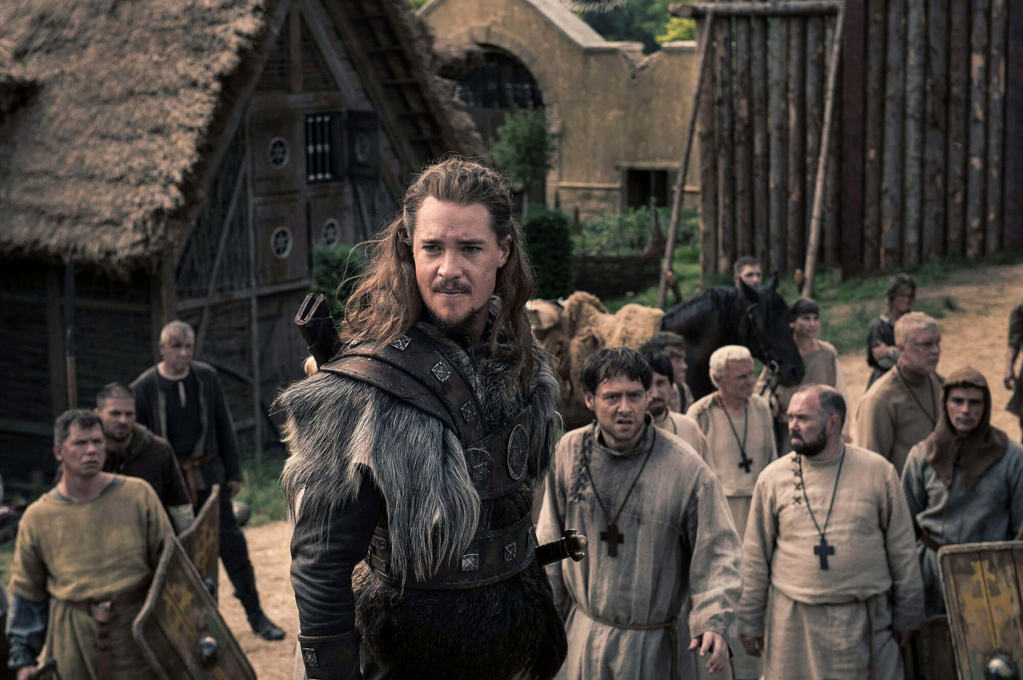 the last kingdom 桌布 壁紙 — 一位長髮、身穿毛皮邊飾盔甲的維京戰士，在中世紀村莊中挑釁地站立，村民和僧侶在旁觀看。