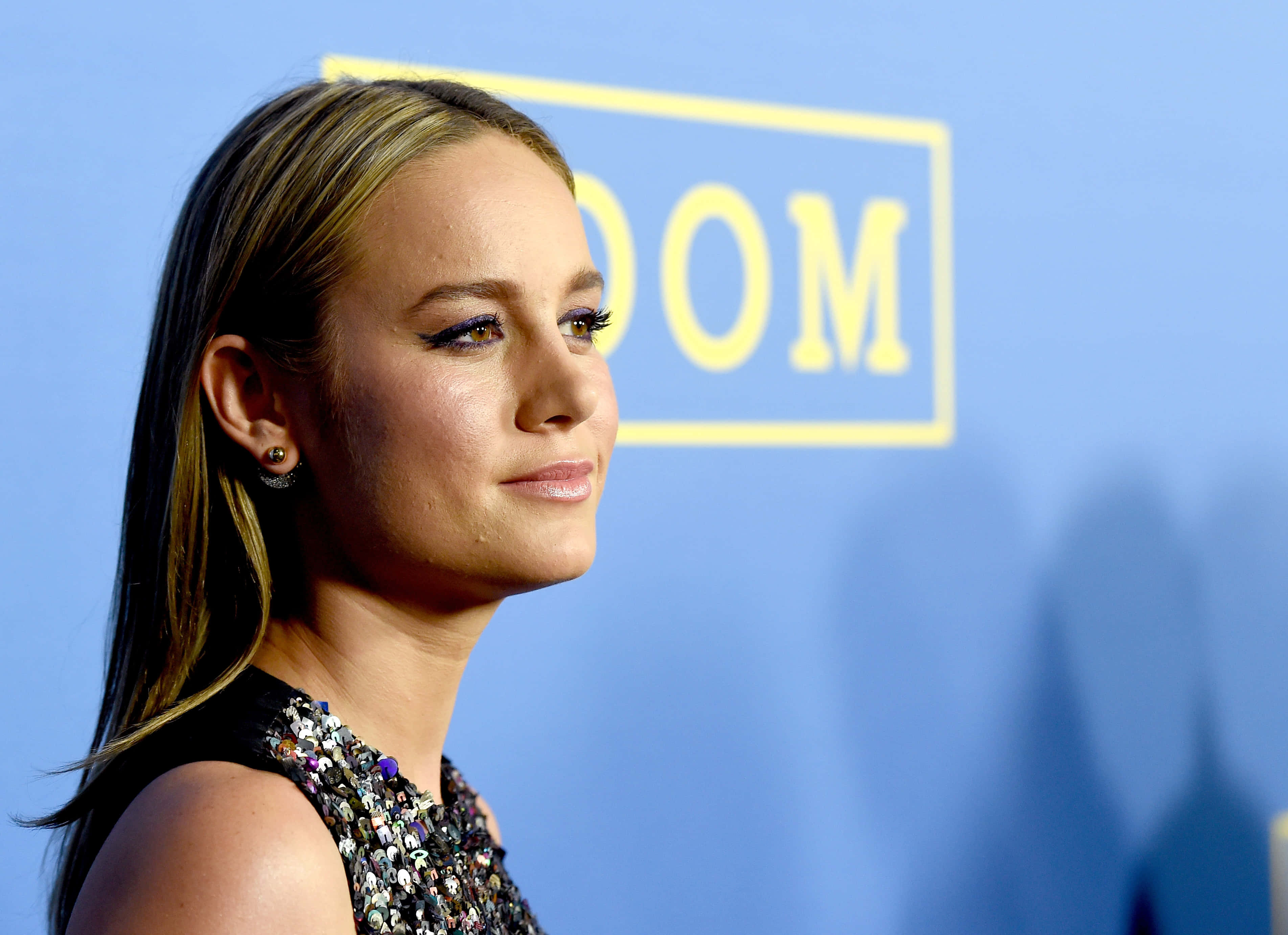 brie larson 桌布 壁紙 — 一位長髮女子身穿閃亮連衣裙，在藍色背景和黃色文字前溫柔微笑。
