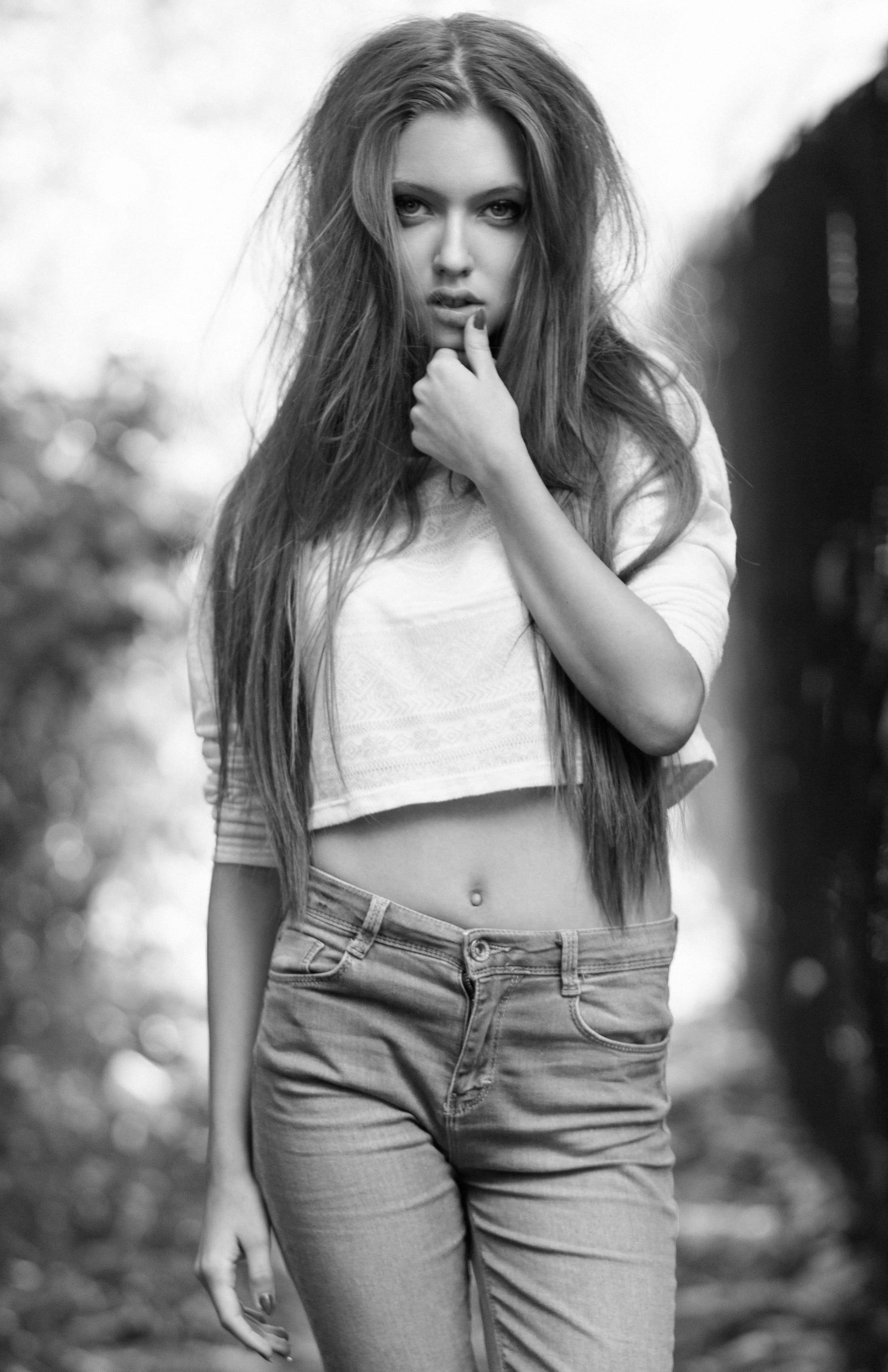 sexy girls 桌布 壁紙 — 一位長髮年輕女性在戶外黑白背景下，手指放在唇邊，若有所思地站著，身穿露臍上衣和牛仔褲。