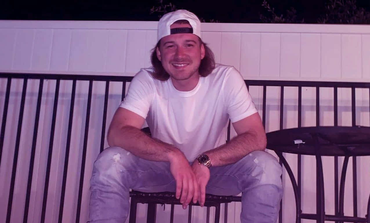 Morgan Wallen 桌布 壁紙 — 一位長髮微笑的男子在紫色夜燈下，隨意地坐在白色籬笆前的黑色金屬長椅上。