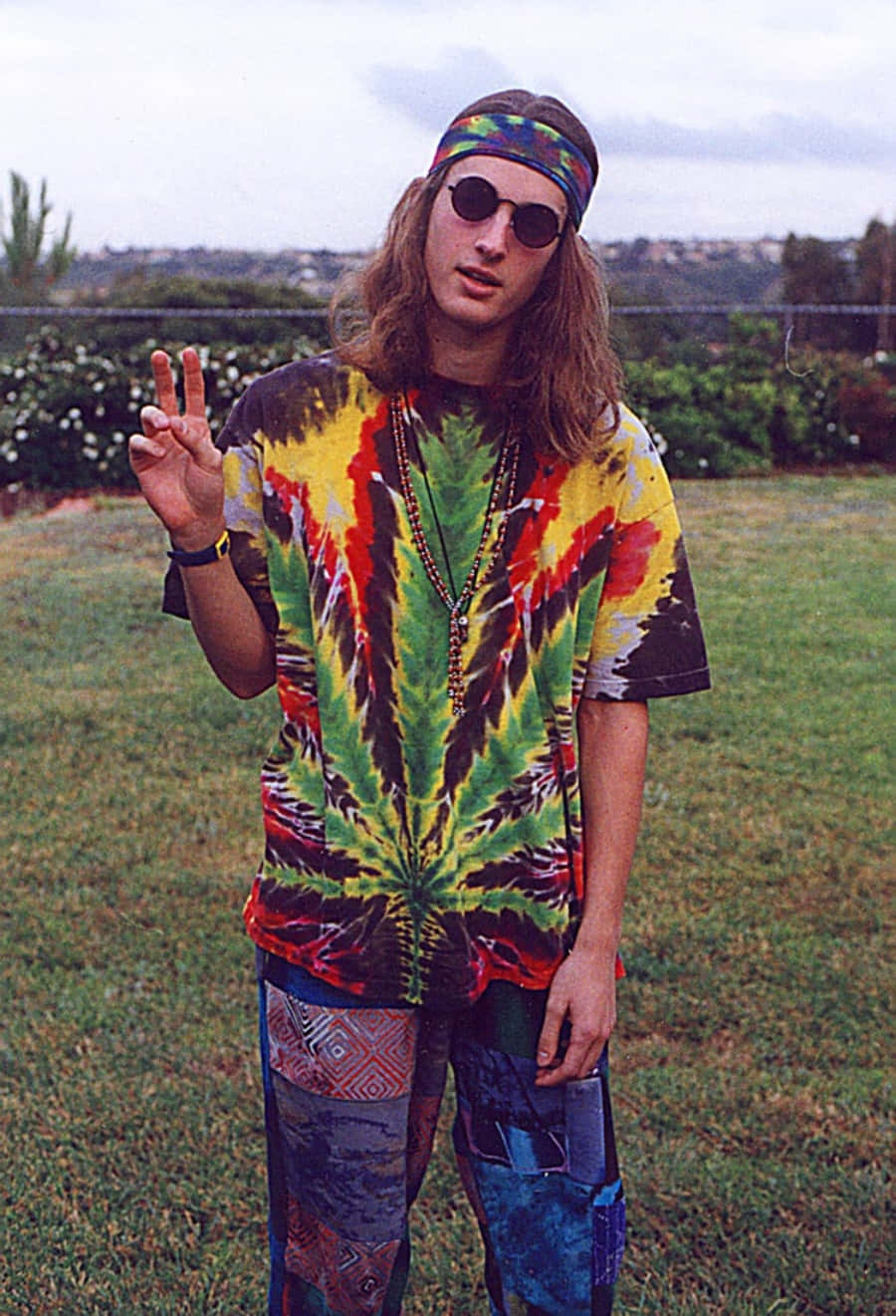嬉皮士 桌布 壁紙 — 一位長髮戴太陽眼鏡的年輕男子穿著鮮豔的紮染襯衫和圖案褲子，在戶外比出和平手勢。