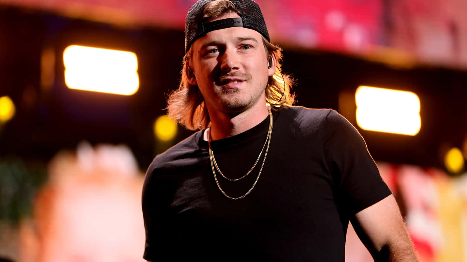 Morgan Wallen 桌布 壁紙 — 一位長髮戴黑色帽子的男表演者在燈光明亮的舞台上自信微笑，身後有發光的燈光。