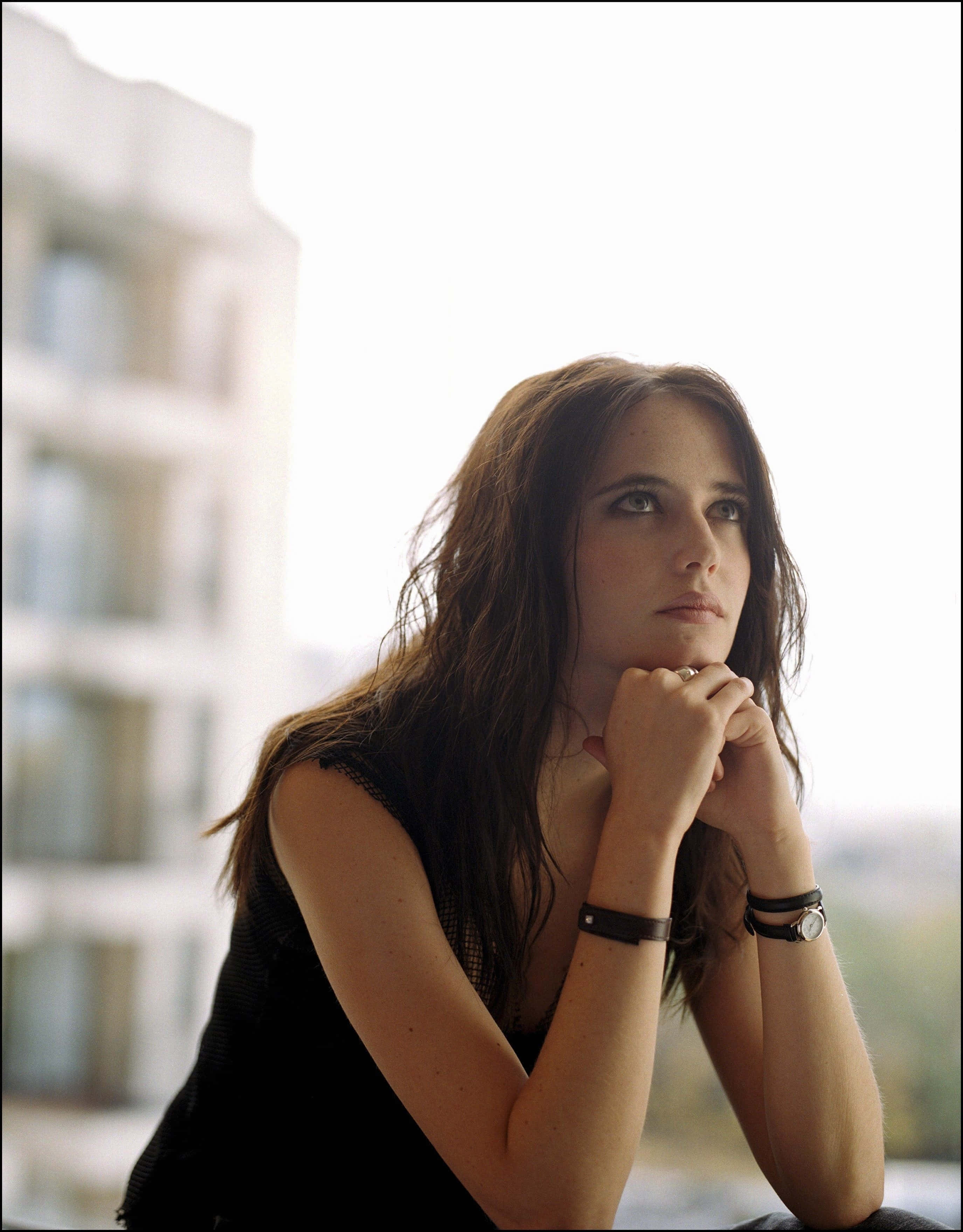 eva green 桌布 壁紙 — 一位長髮棕眼女性將下巴擱在交疊的雙手上，對著明亮的城市背景若有所思地向上凝視。