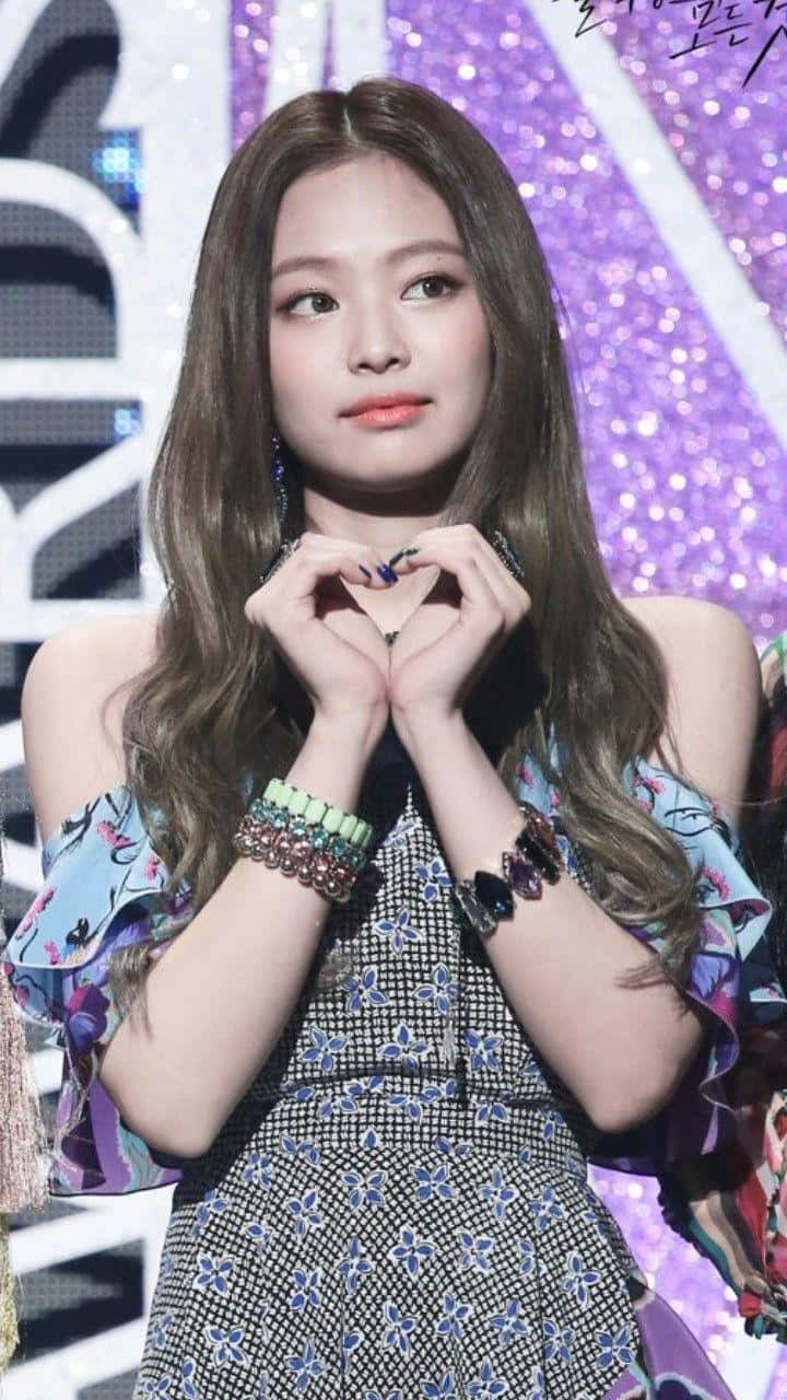 Jennie Kim 桌布 壁紙 — 一位長髮棕色年輕女性穿著花卉連衣裙，在閃爍的紫色背景下用手做出心形。