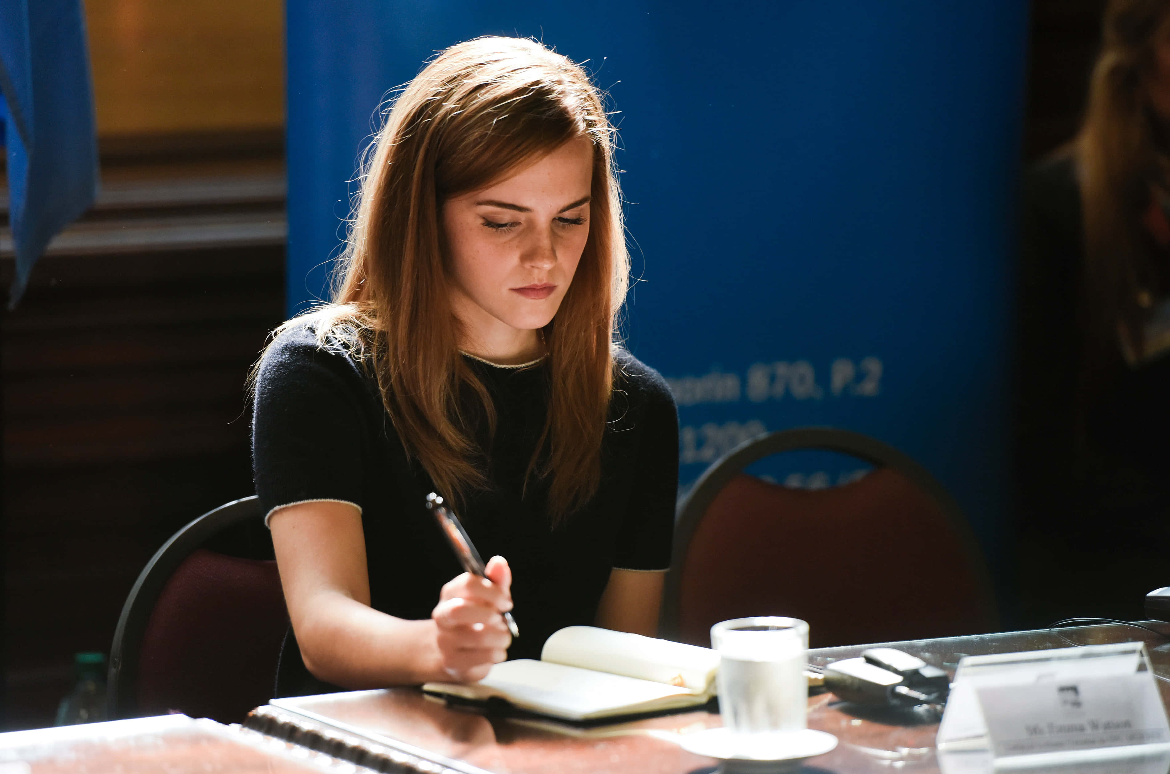 Emma Watson 桌布 壁紙 — 一位長髮棕色的年輕女子在溫暖的陽光下於木桌上寫筆記。