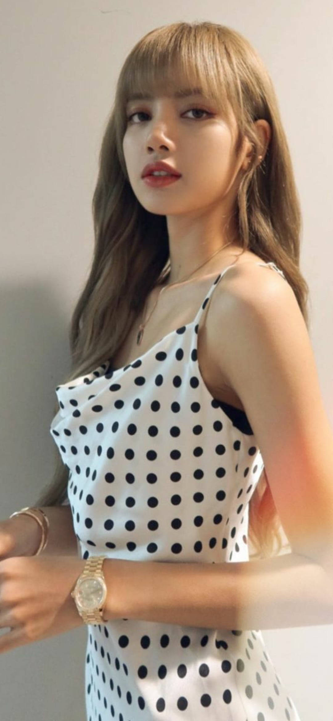 lisa 桌布 壁紙 — 一位長髮棕色的年輕女性，身穿白色波點連衣裙，佩戴金色飾品，背景是素色牆壁。
