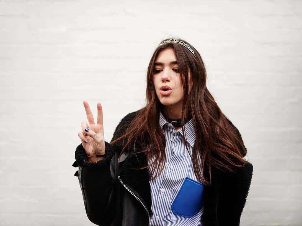 Dua Lipa 桌布 壁紙 — 一位長髮棕色的年輕女性在純白磚牆前做出和平手勢和調皮表情。