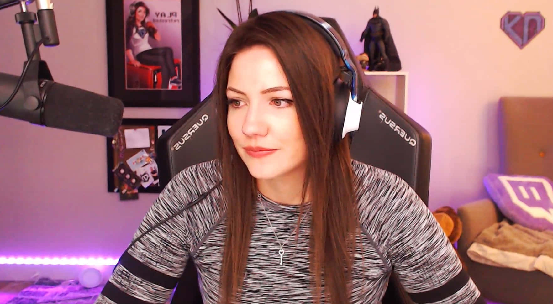 Minecraft Twitch Streamers 桌布 壁紙 — 一位長髮棕色的年輕女性坐在黑色電競椅上，戴著耳機，穿著灰色圖案襯衫，在一個柔和燈光的房間裡，配有紫色LED燈和Twitch主題裝飾。