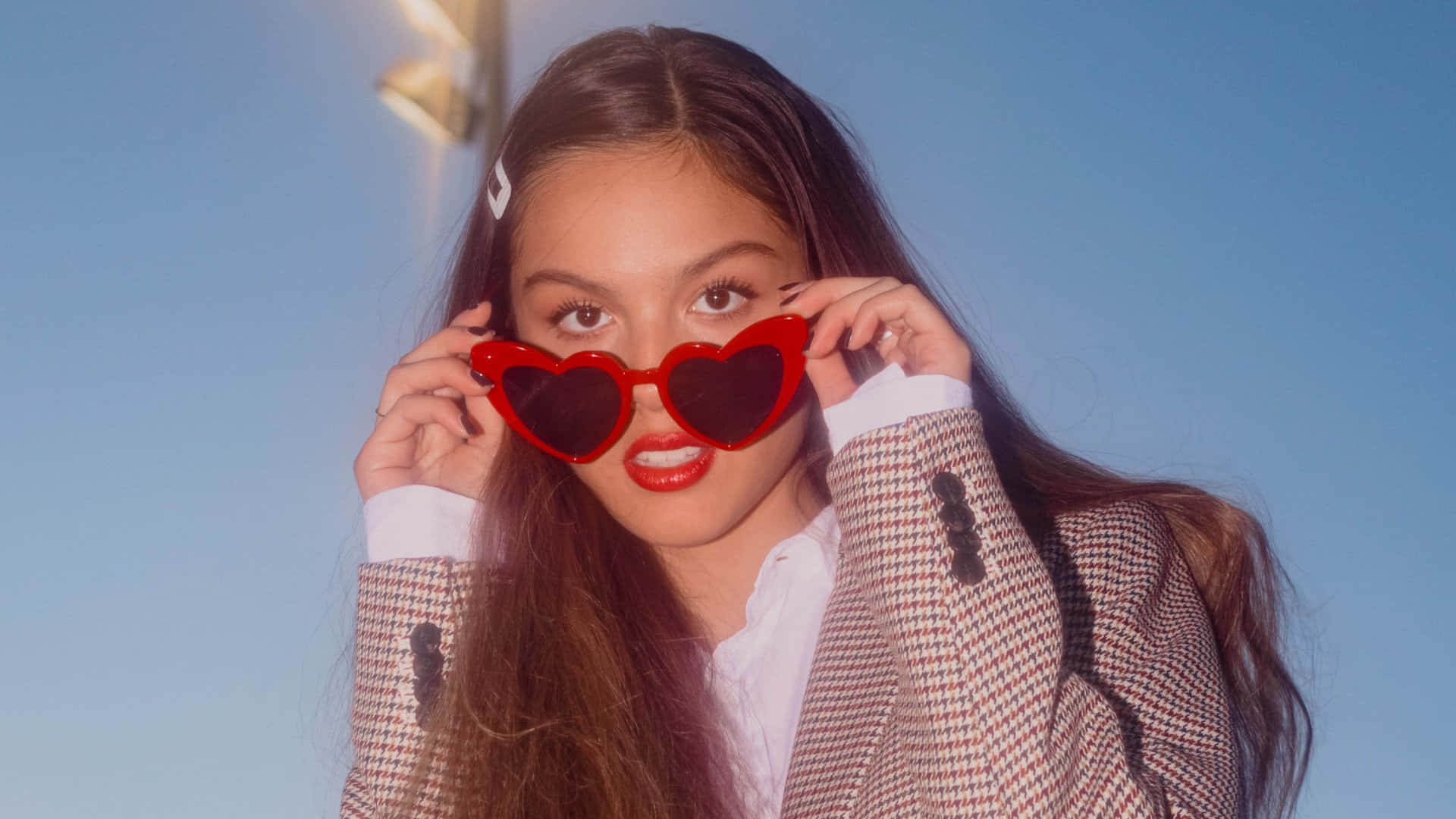 Olivia Rodrigo 桌布 壁紙 — 一位長髮棕色的年輕女性戴著大膽的紅色心形太陽眼鏡，背景是清澈的藍天。