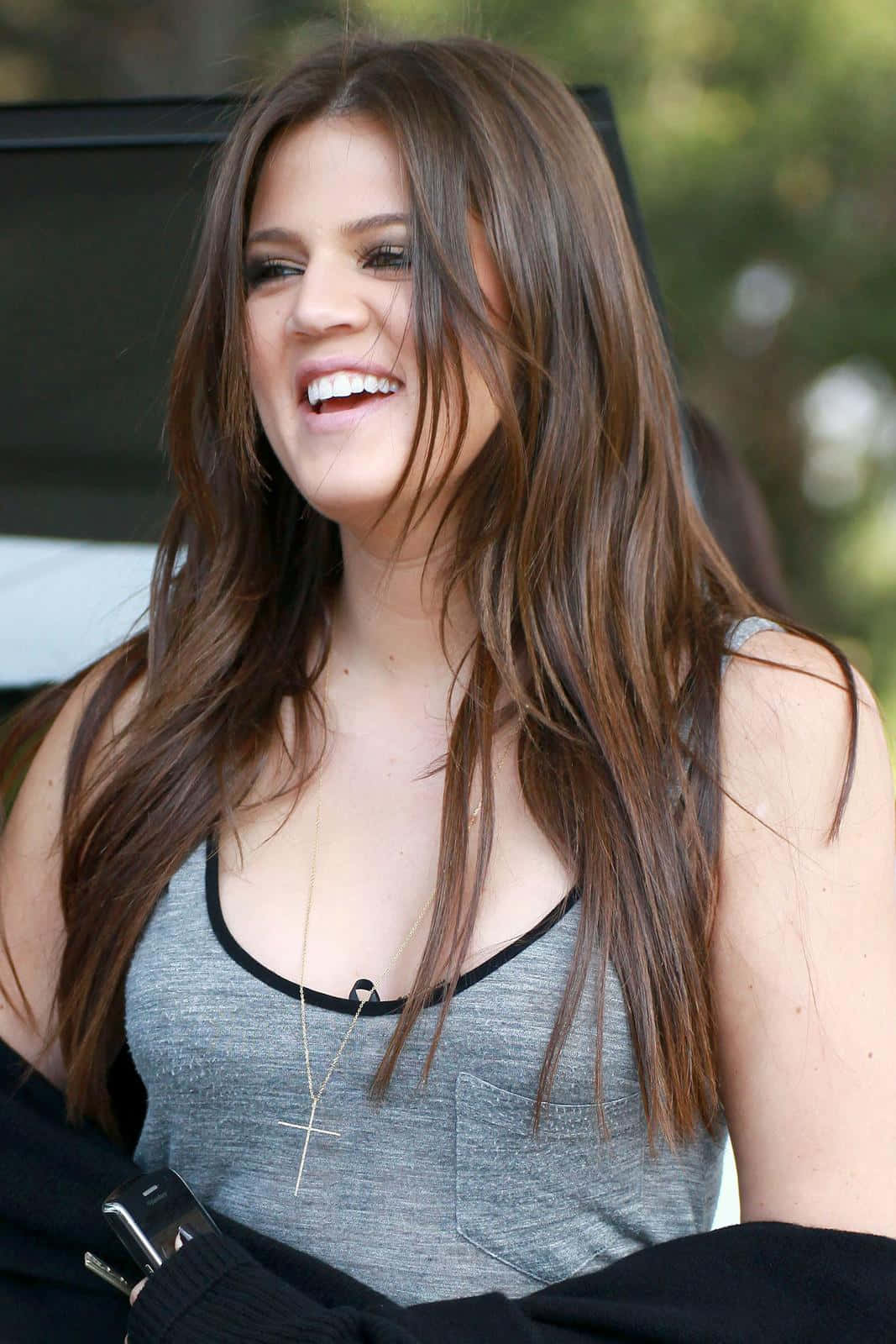 Khloe Kardashian 桌布 壁紙 — 一位長髮棕髮女性，穿著灰色背心和十字架項鍊，開懷大笑。