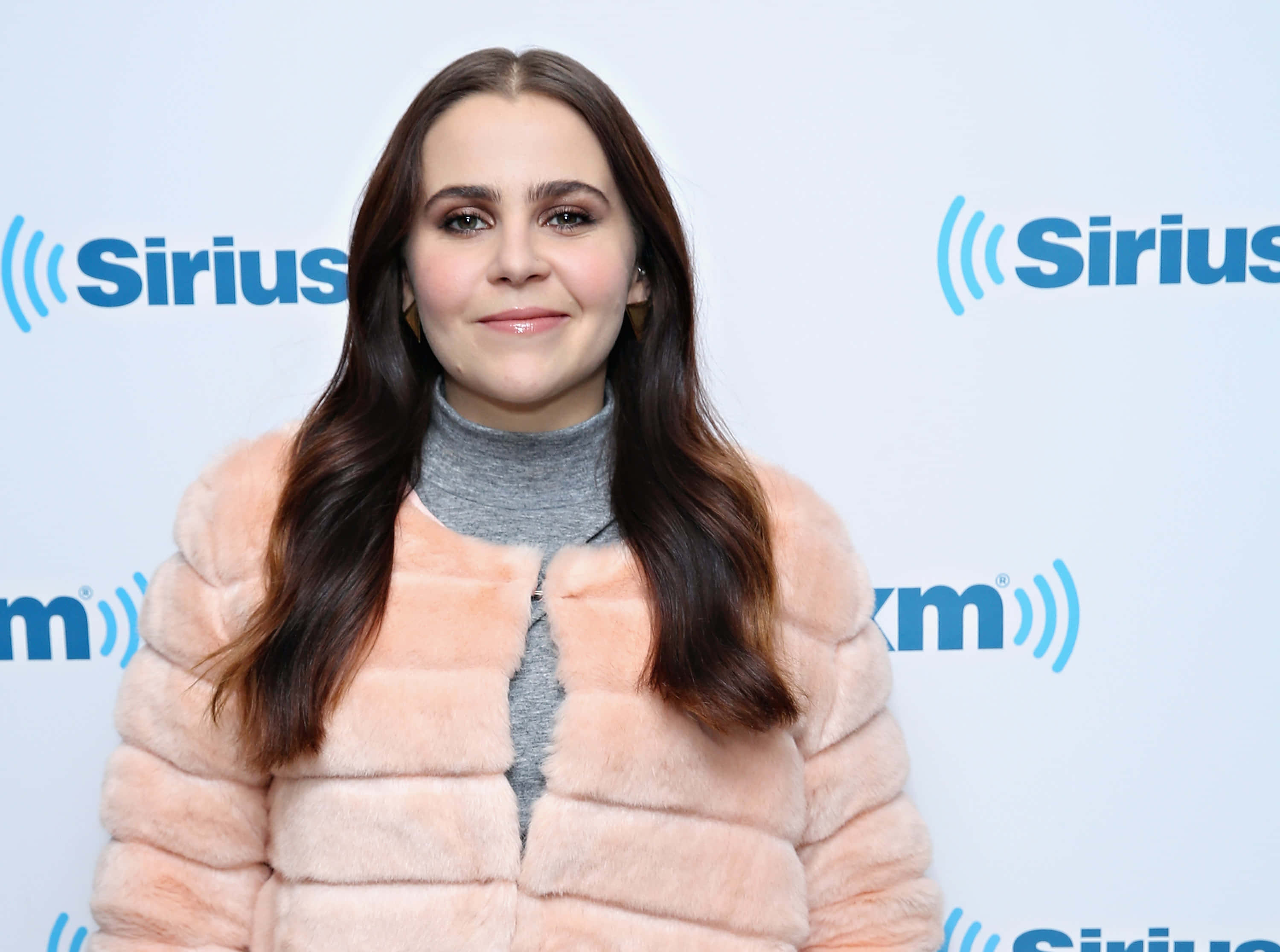 Mae Whitman 桌布 壁紙 — 一位長髮棕髮女性微笑著，身穿桃色皮草外套，內搭灰色高領毛衣，背景為SiriusXM品牌標誌。