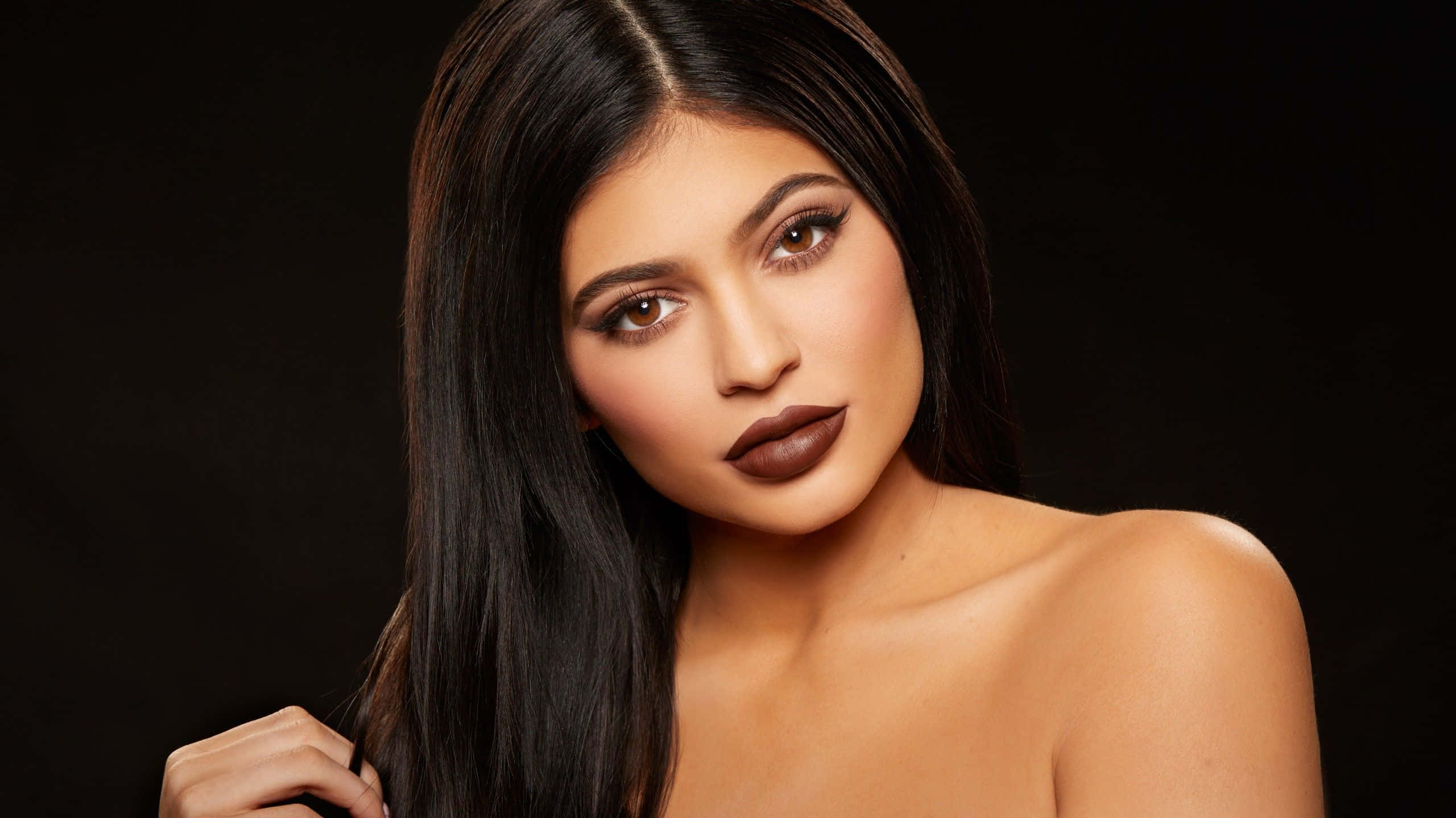 Kylie Jenner 桌布 壁紙 — 一位長髮深色、塗著大膽棕色唇膏的女性在黑色背景前的迷人肖像。