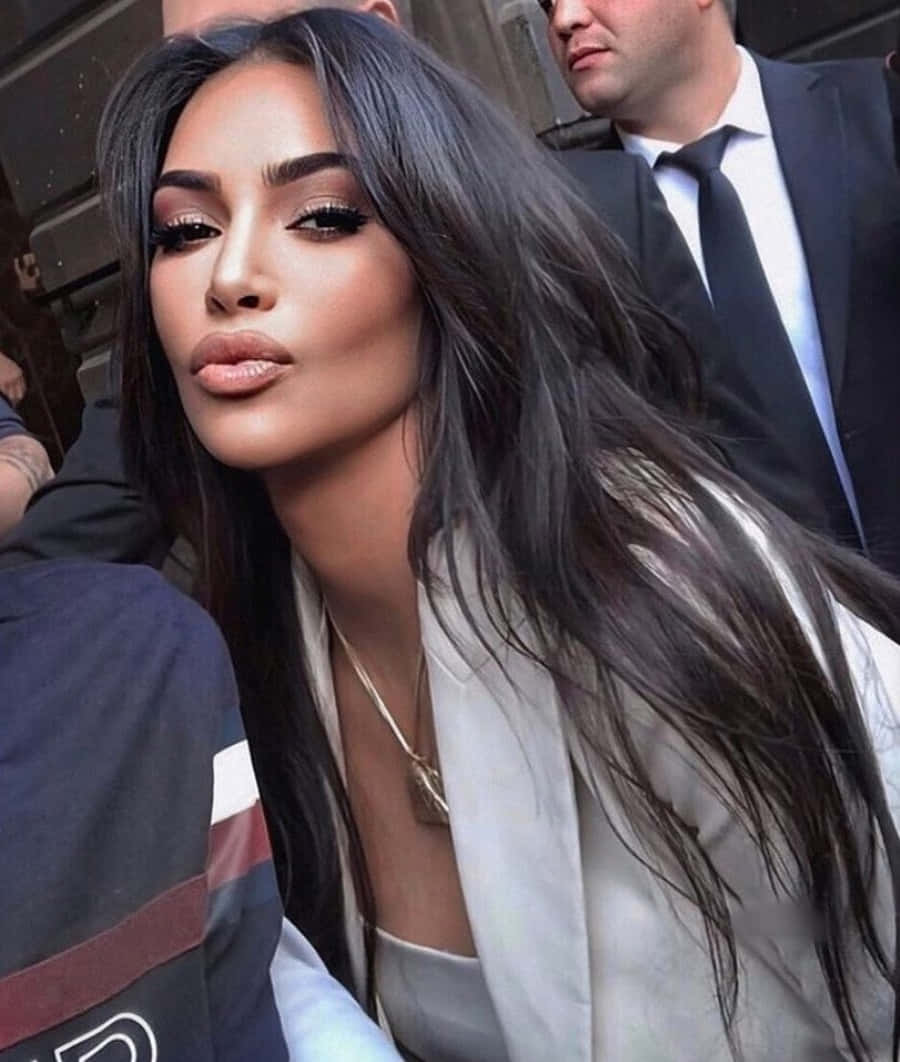 Kim Kardashian 桌布 壁紙 — 一位長髮深色、妝容大膽的迷人女性，在人群中自信地穿著白色西裝外套擺姿勢。